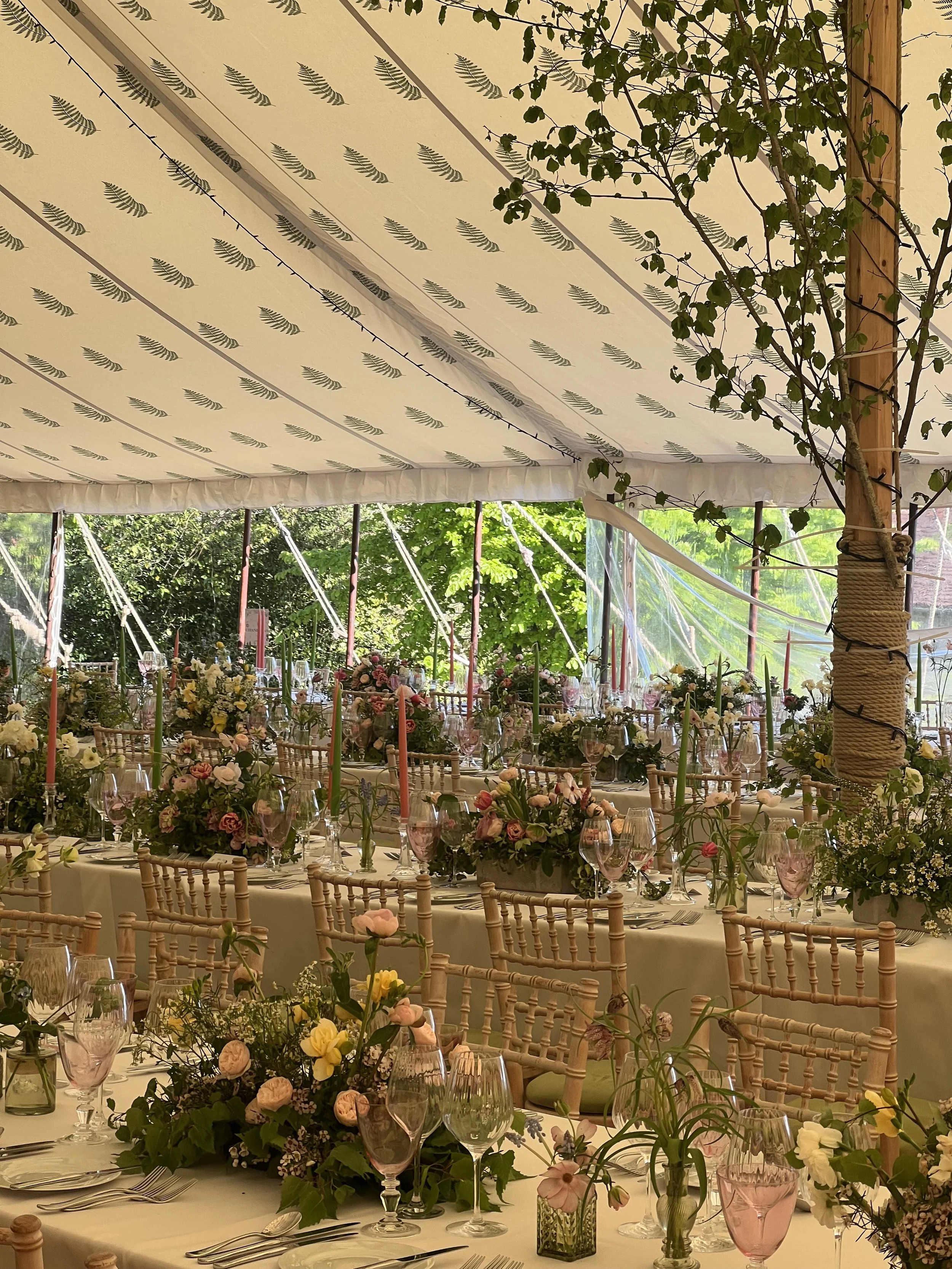 A Luxuriously decorated banquet table set for a wedding or celebration with floral centerpieces, wine glasses, and chairs in a tent with a floral pattern ceiling and surrounding greenery.