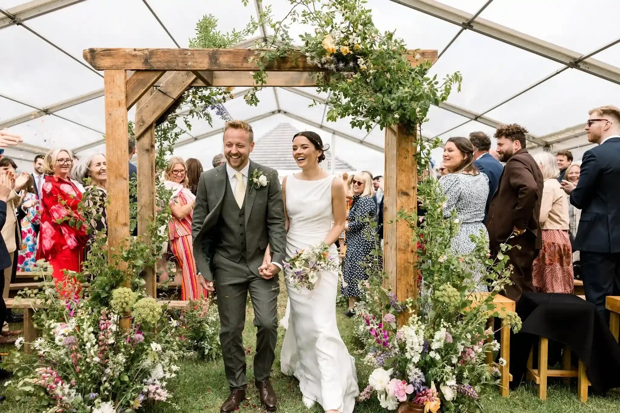 A newly married couple coming through their beautiful rustic floral arch created by Alex Ball from Flowers in Rye, at the Cherry Barn wedding venue in June