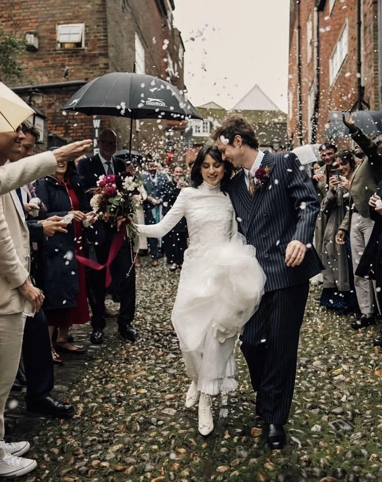 Bride and groom walking together under umbrellas while guests celebrate and throw rice or confetti at a wedding outdoors, a wedding in Rye, East Sussex