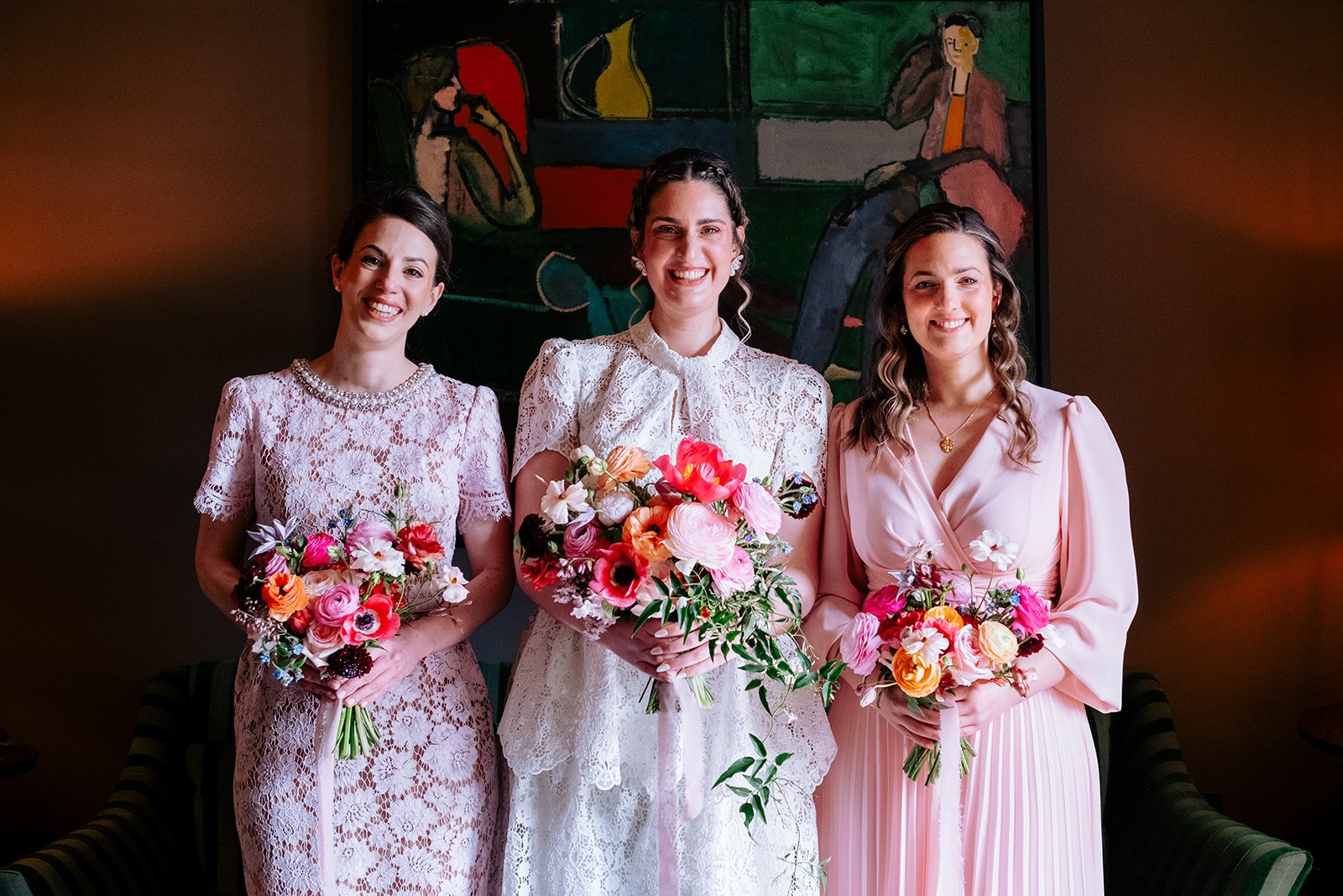 A Bride and her two bridesmaids in elegant lace and silk gowns, holding colourful bouquets of spring flowers, standing in front of a dark background with a modern painting, smiling at the camera at the George in Rye