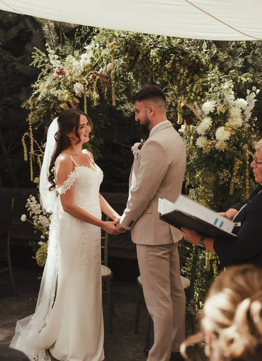 This photo is of a bride and groom taking their wedding vows in the Courtyard at The George in Rye, standing infront of a beautiful floral arch, made by Alex Ball Wedding Florist in Rye East Sussex.  its high summer and the flowers are seasonal