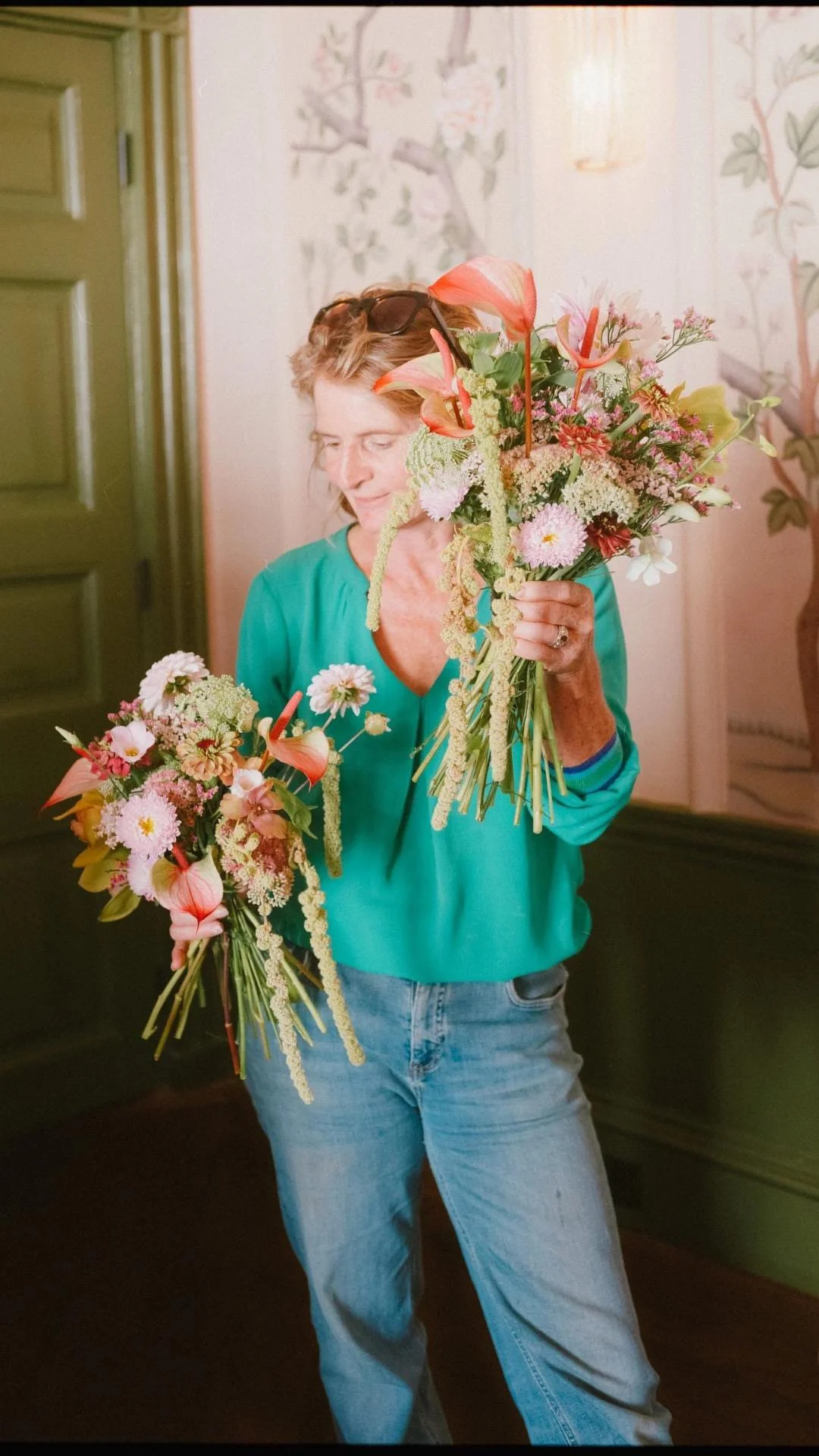 Lead Florist Alex Ball holding two large bouquets of pink, green, and white flowers, in a modern style, the picture is  inside the ballroom at the George Hotel in Rye,  with walls decorated with floral wallpaper.