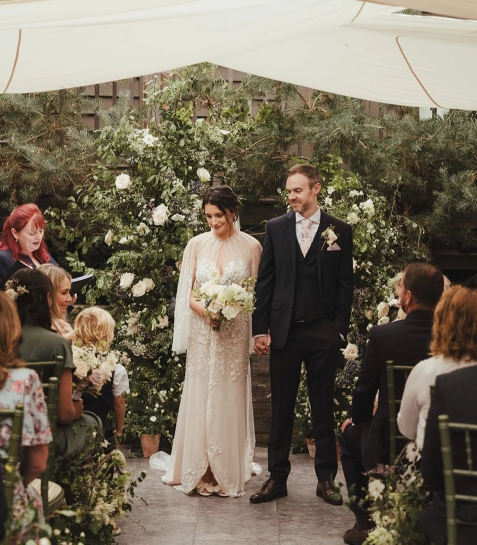 A bride and groom holding hands during their wedding ceremony, standing in front of a floral arch with guests seated on either side, outdoors under a canopy in the Courtyard at the George in Rye.