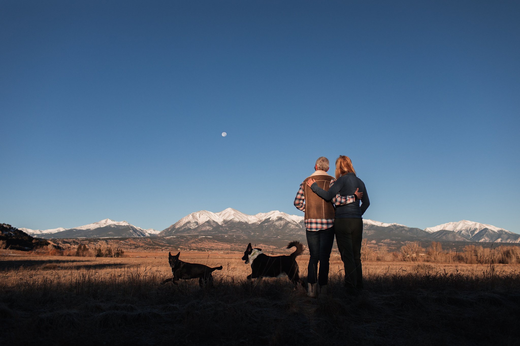 A silhouetted couple stands facing a field and the snow-capped Collegiate Peaks while their dogs run playfully and the full moon dots the blue sky