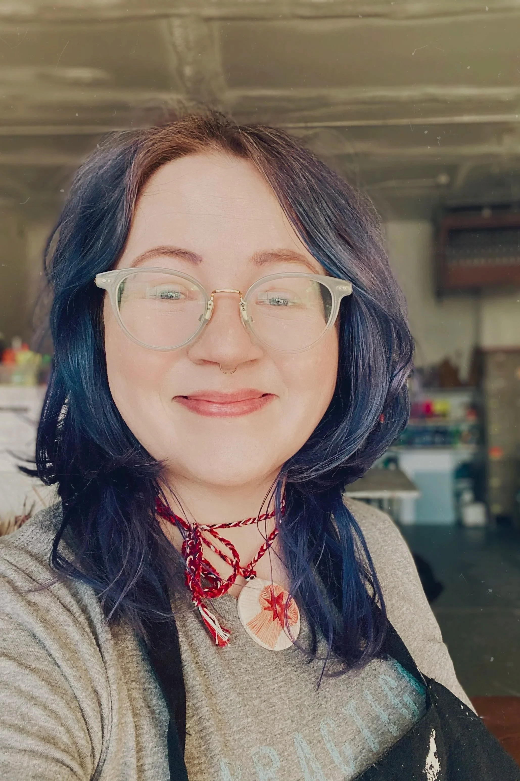 Picture of the artist, a white femme with glasses and deep blue hair wearing a handmade necklace and smiling in the studio.