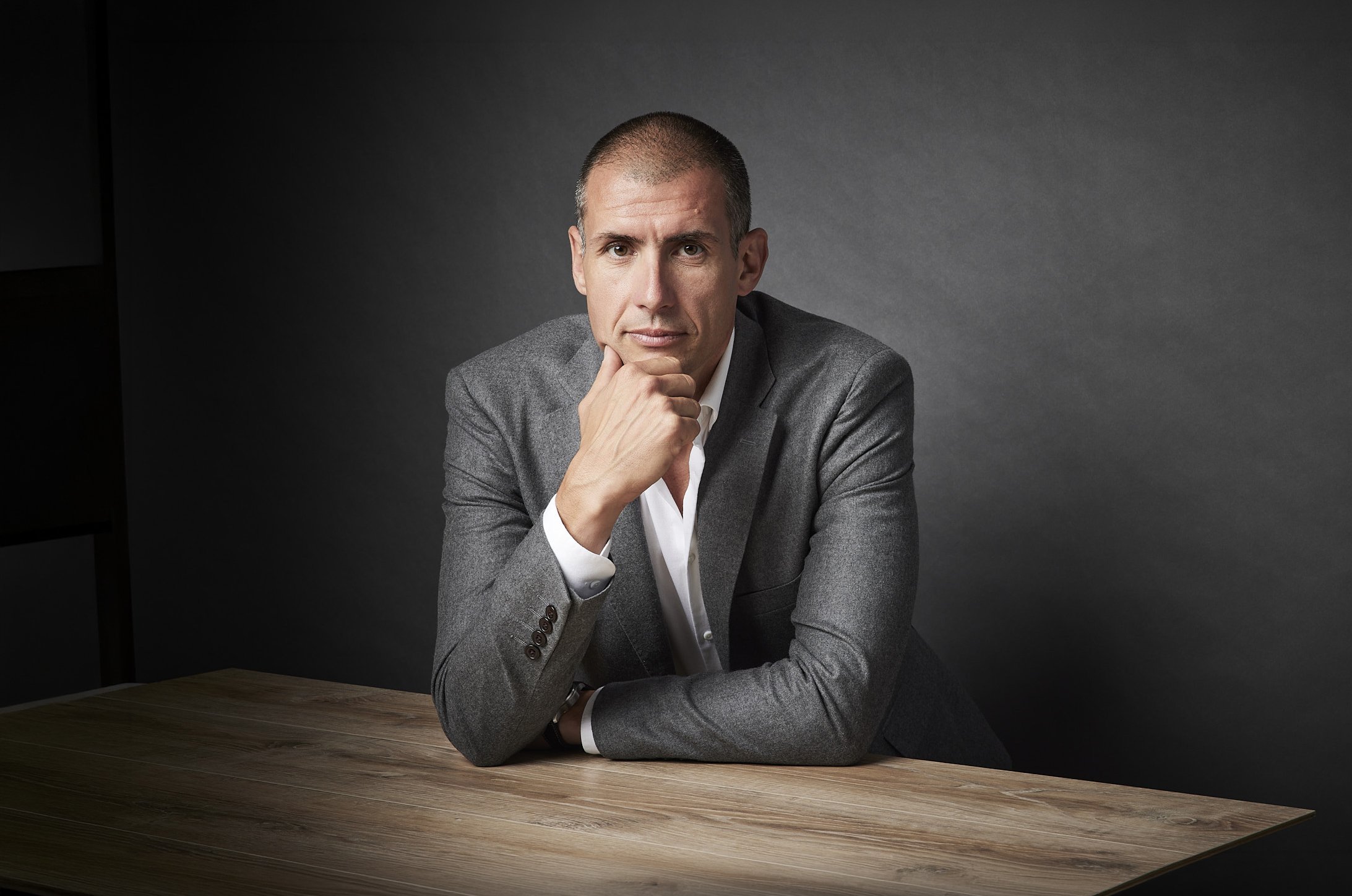 Professional corporate portrait of a man in a grey suit seated at a table against a dark background.