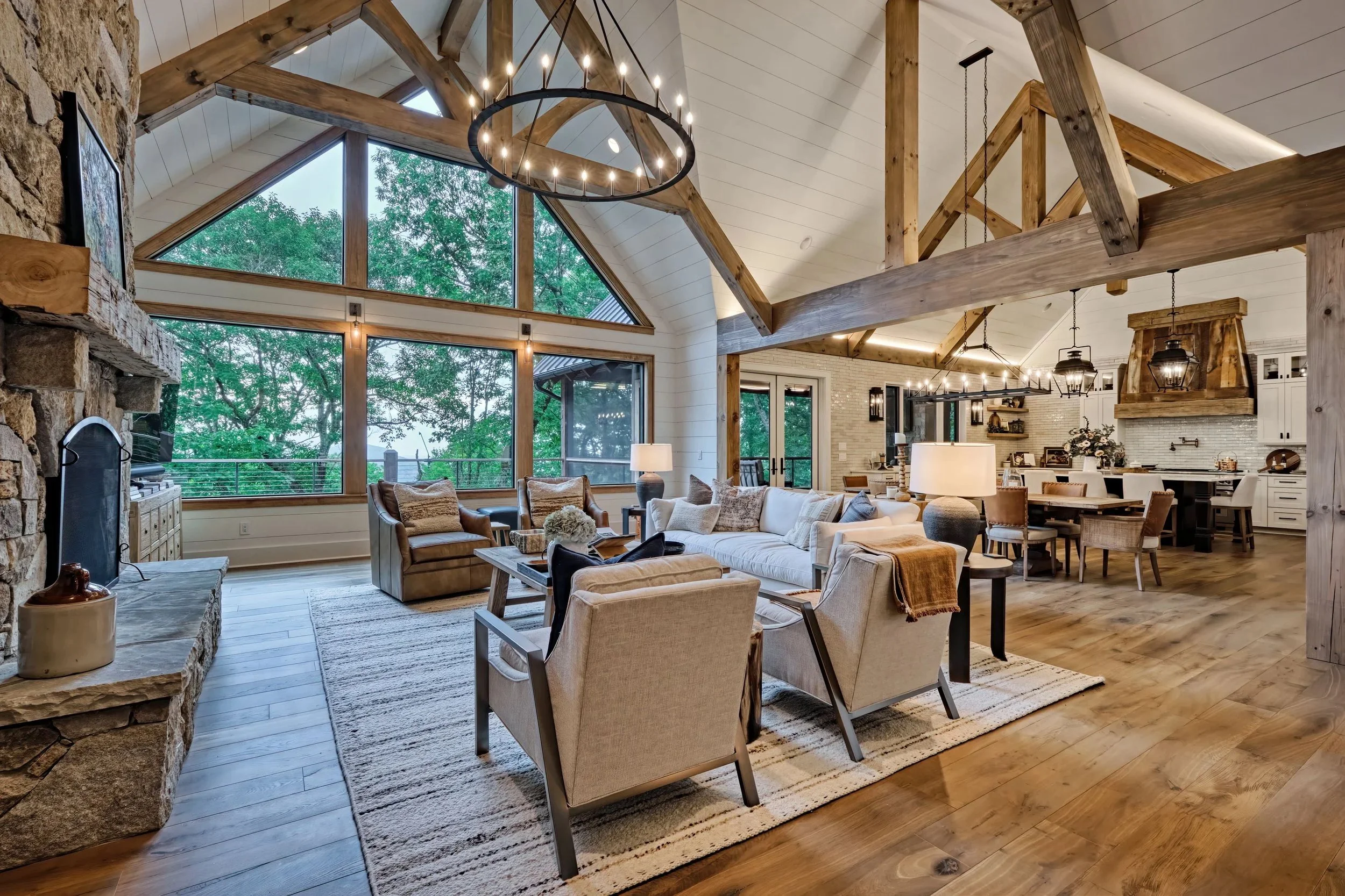 A spacious open-concept living room and kitchen with high vaulted ceilings, large windows, wooden beams, and rustic decor. The living area has a beige sofa, armchairs, lamps, and a stone fireplace, overlooking a tree-filled view outside.