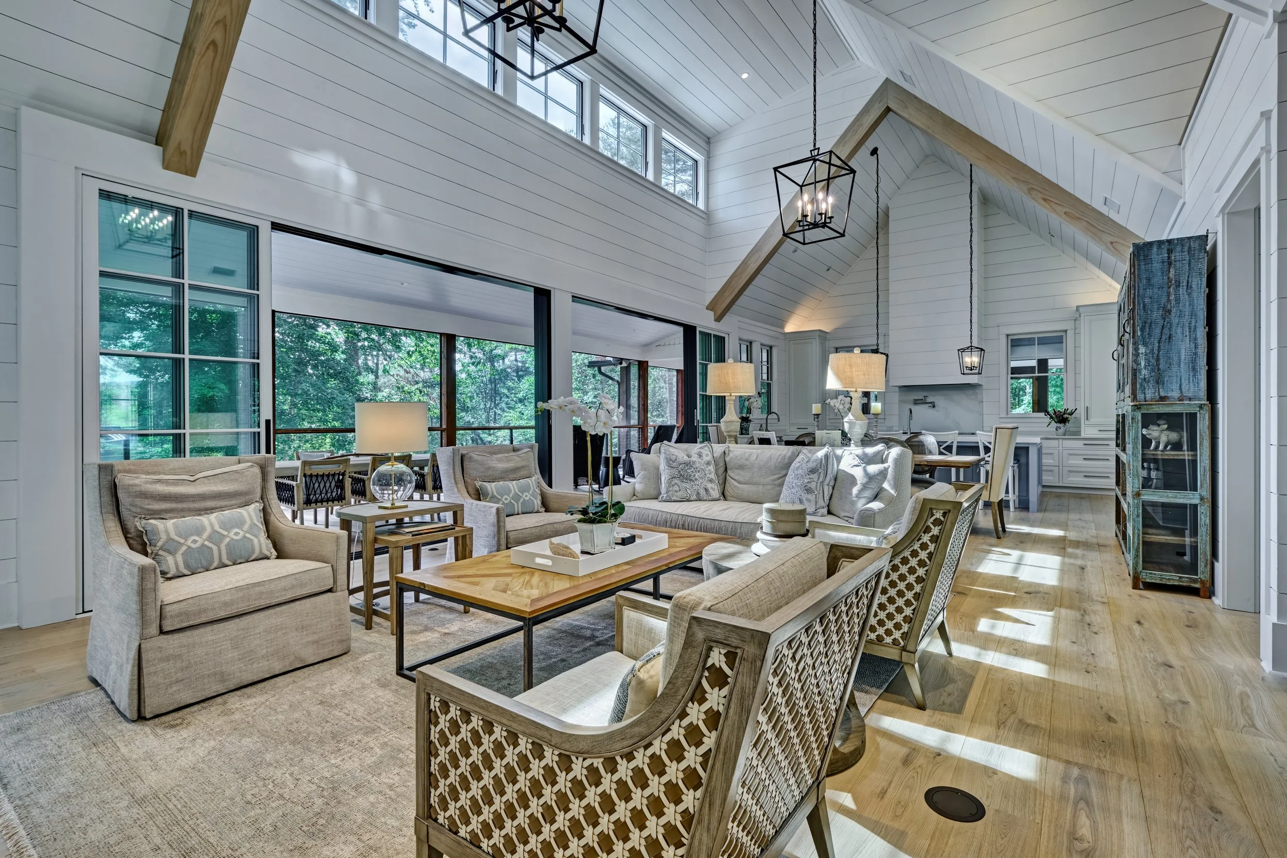 Living room with white shiplap walls, high vaulted ceiling, large windows, and hardwood floors. Furniture includes beige sofas and armchairs, a wooden coffee table, and decorative lamps. Natural light fills the space, with views of greenery outside.