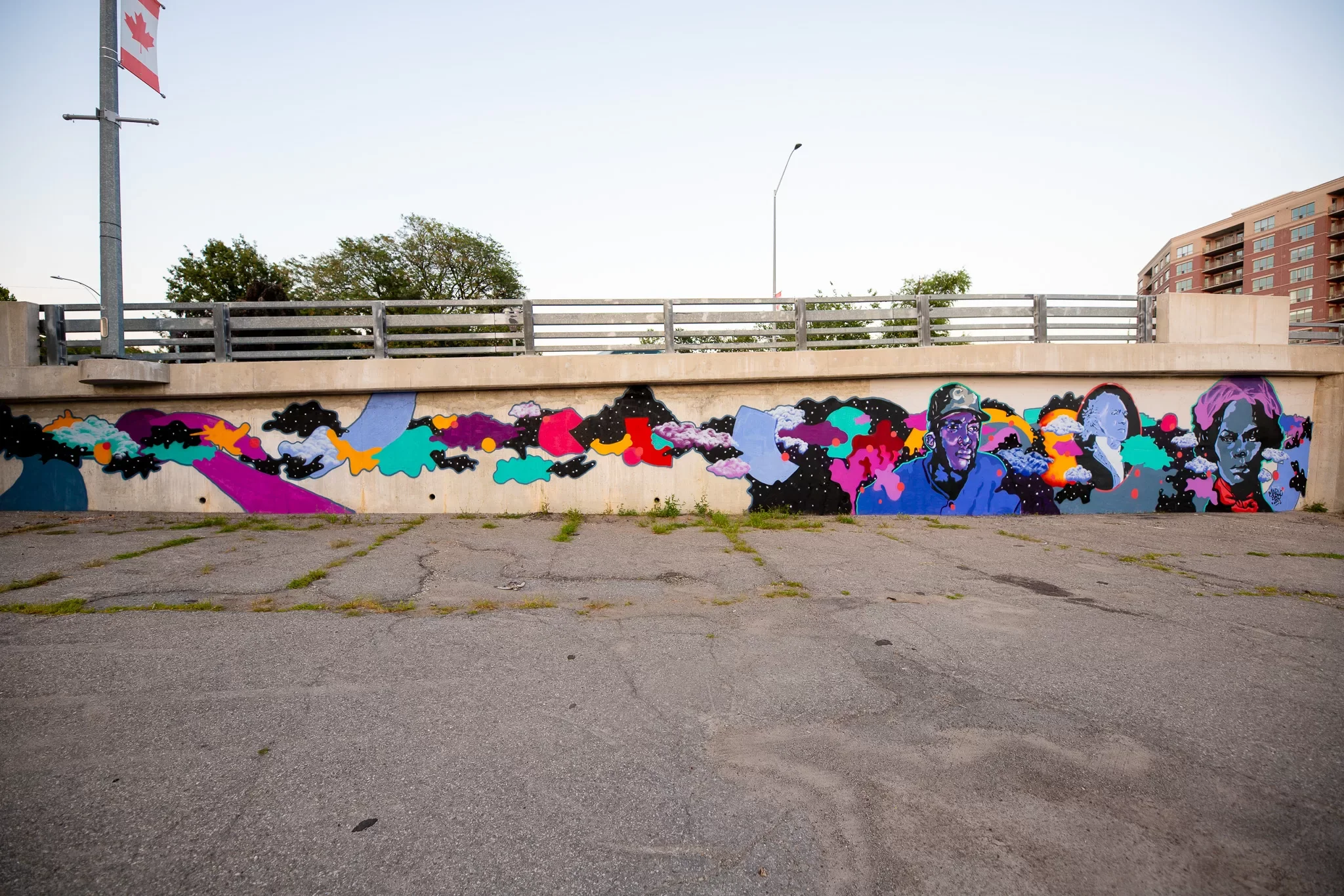 Colorful mural painted on a concrete wall along a street, featuring abstract designs and portraits of people. An overpass with a railing is above the mural, with trees and a multi-story building in the background.
