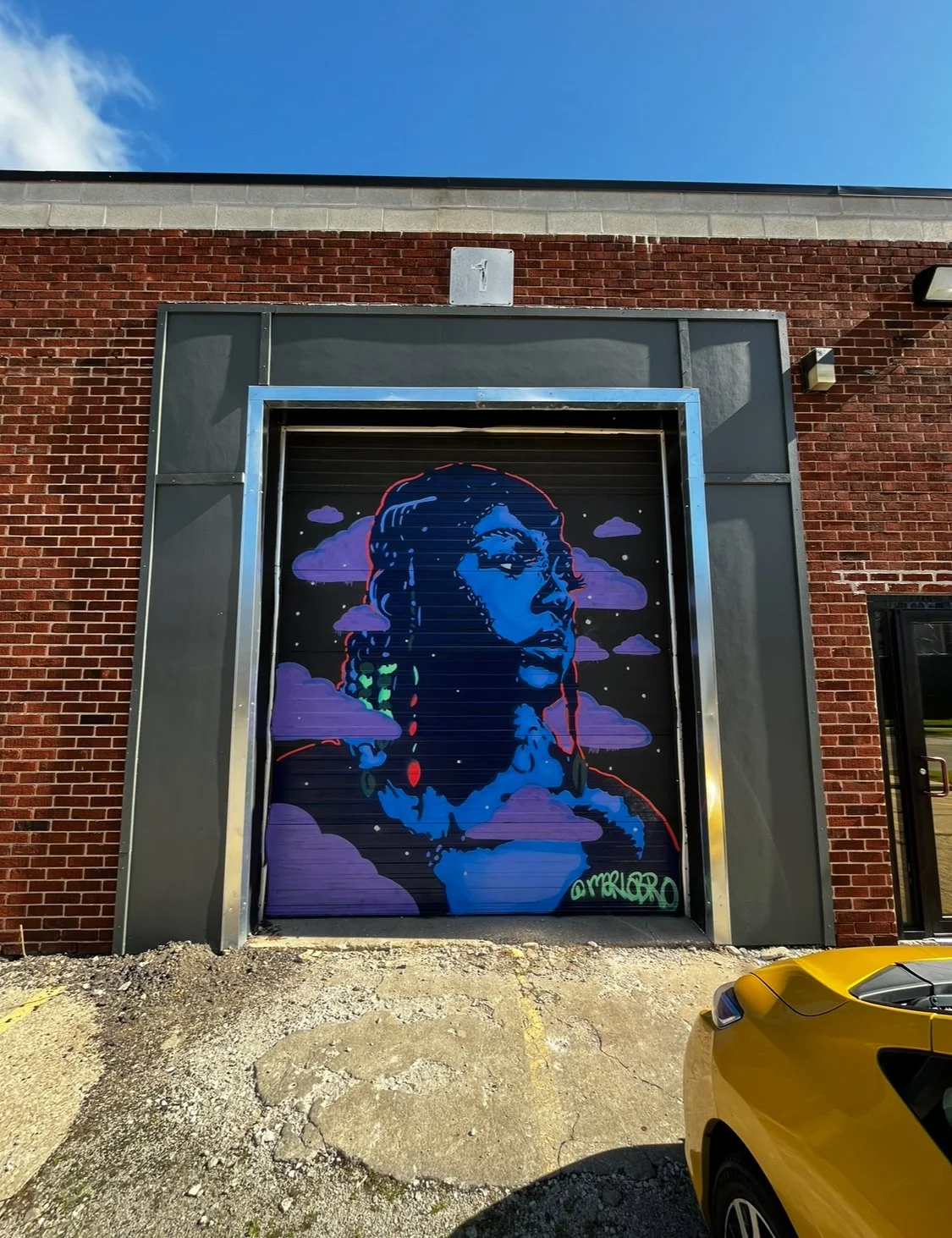 Street art mural of a woman's profile with colorful hair and clouds in the background on a metal garage door, with red brick wall around it.