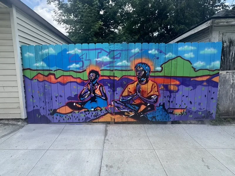 Colorful mural on a wooden fence depicting two people sitting outdoors with their hands in a prayer position, mountains, purple fields, and a partly cloudy sky in the background.
