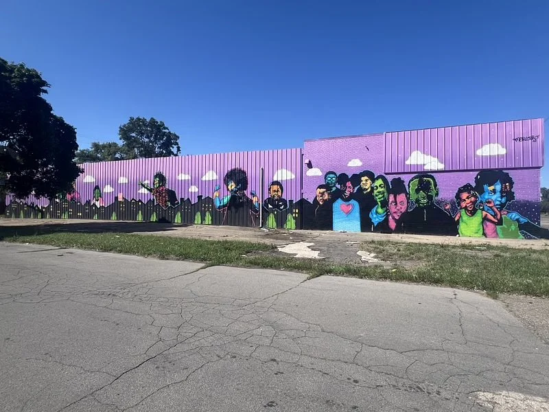 A colorful mural painted on a purple wall features a diverse group of people with a backdrop of purple sky and clouds. The mural is on a large outdoor wall beside a cracked pavement and grassy area.