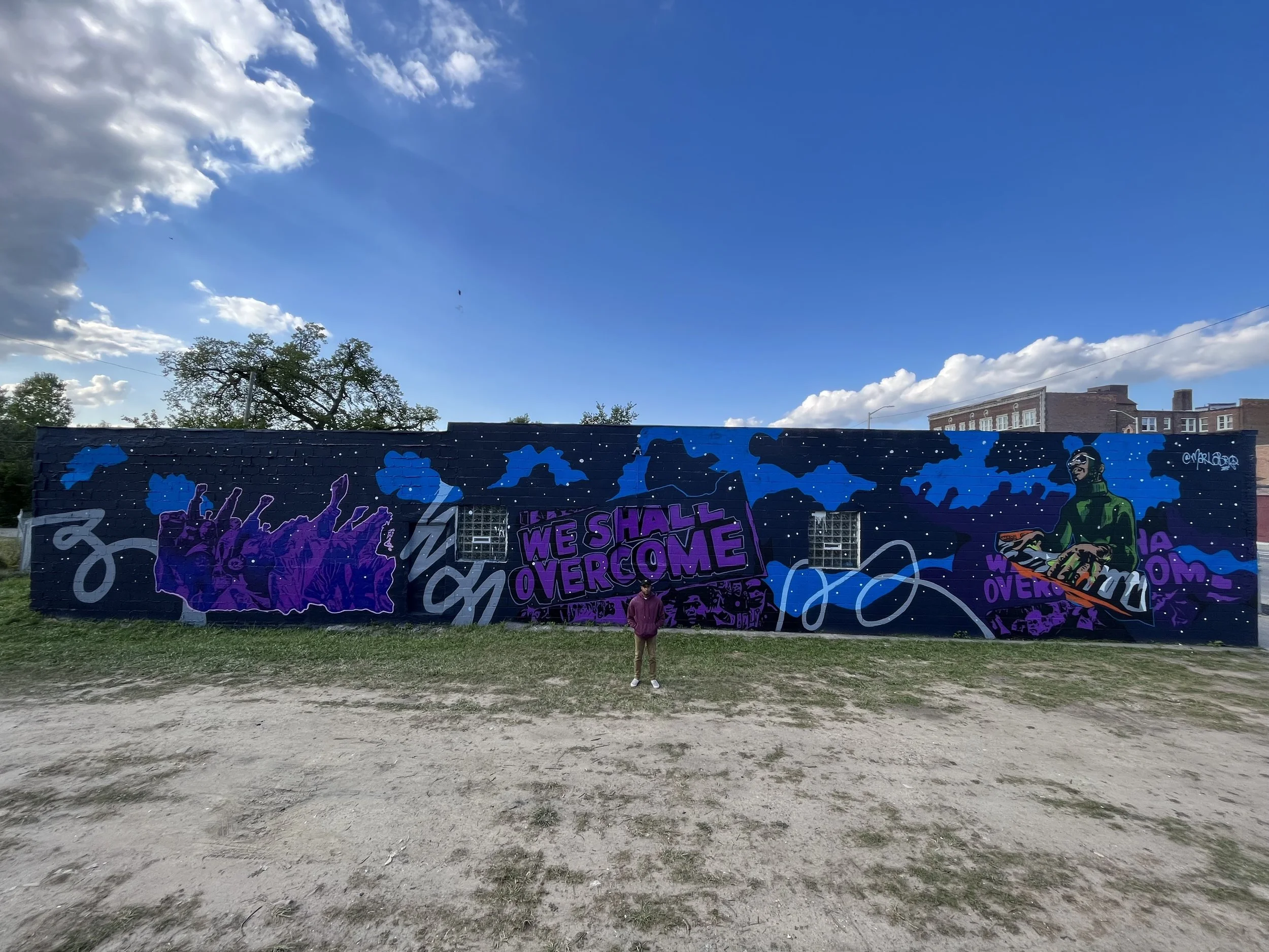 Colorful graffiti mural on a dark blue brick wall with abstract shapes, text, and a person sitting near the wall, with a clear blue sky and some buildings in the background.