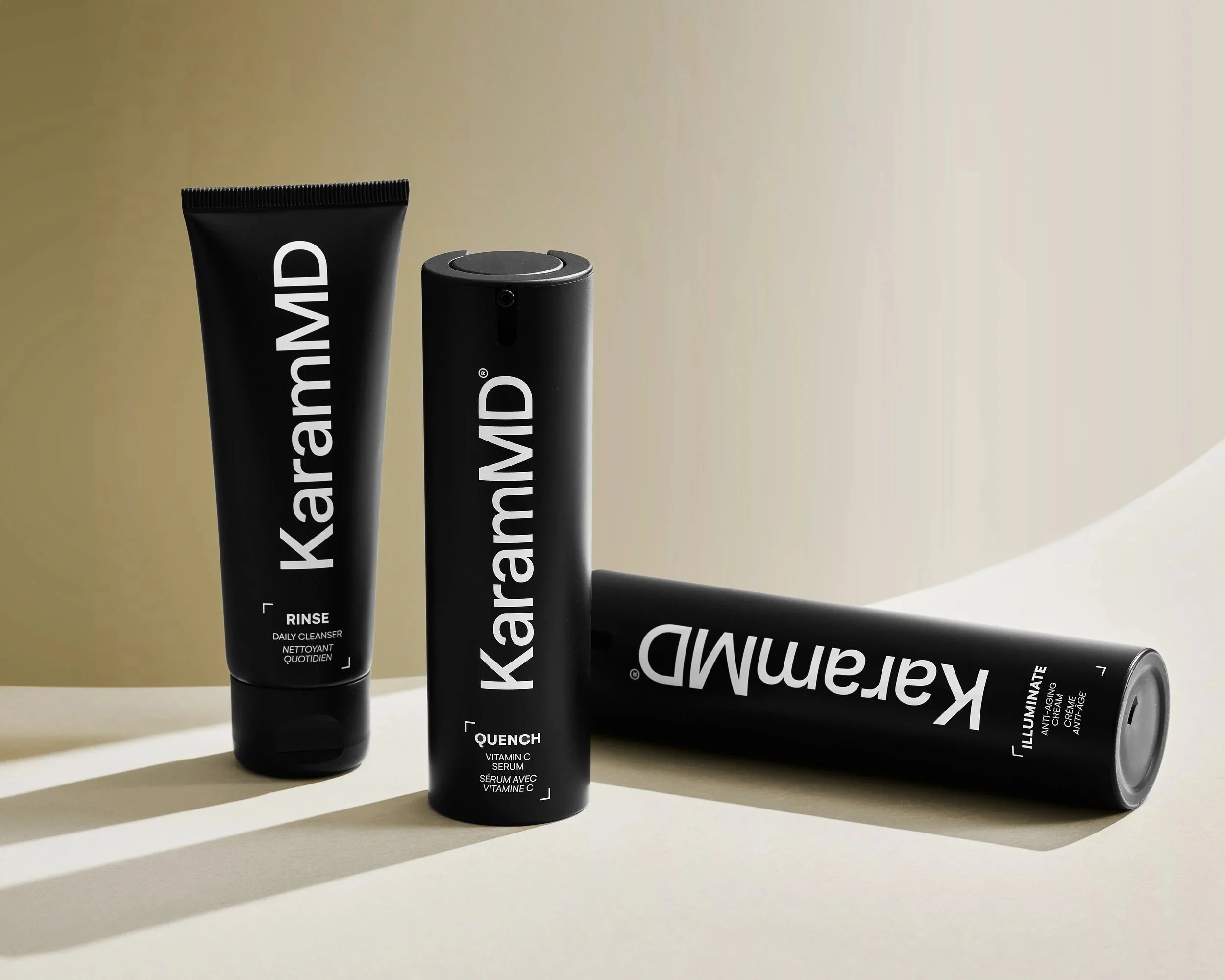 Professional studio product photography of three KaramMD skincare containers—Rinse Daily Cleanser, Quench Vitamin C Serum, and Illuminate Anti-Aging Cream—arranged on a neutral tan surface with dramatic directional shadows.