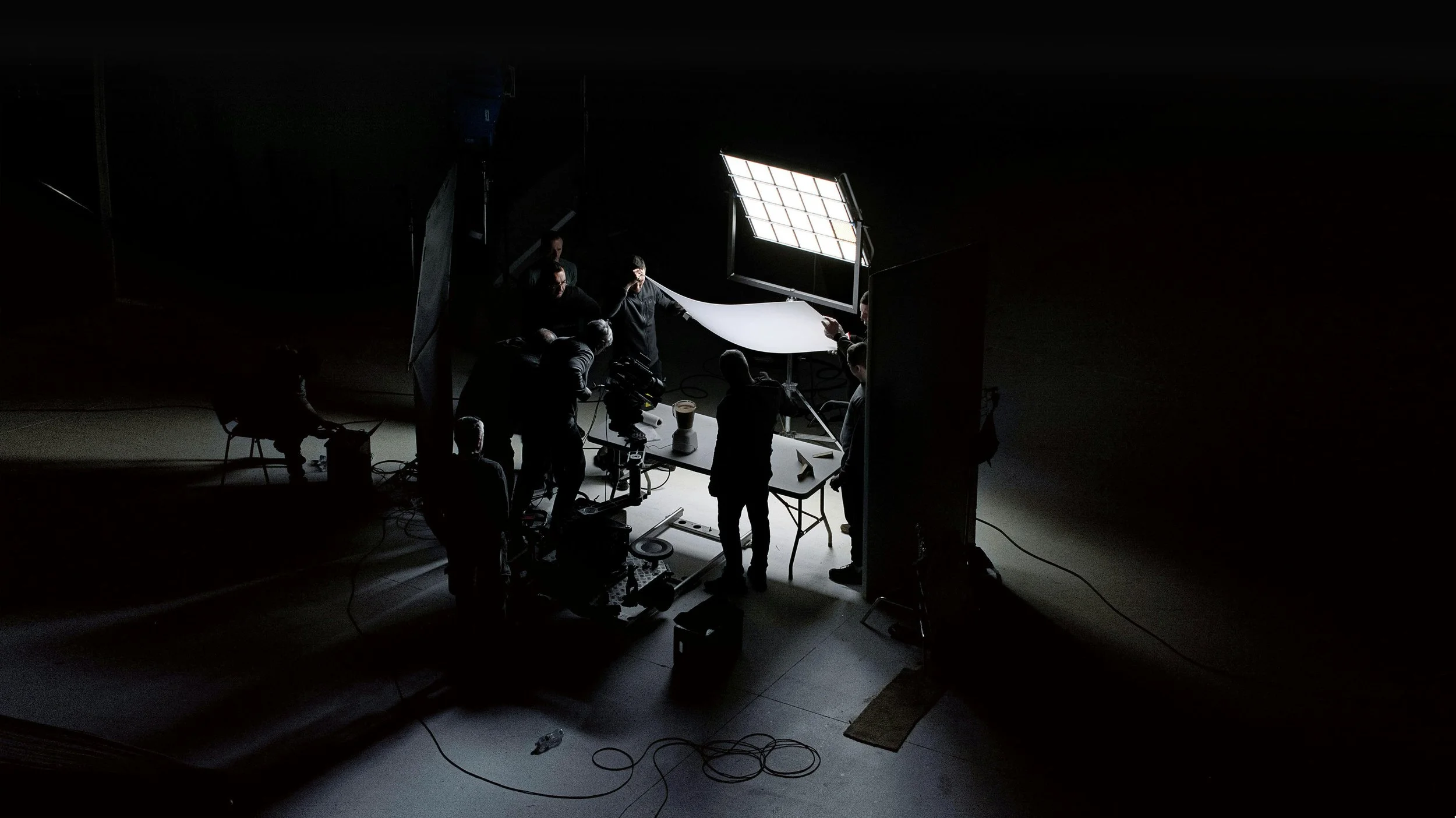 A behind-the-scenes view of a film or photo shoot studio with crew members working around a table with equipment, lighting, and backdrop setup in a dark space.