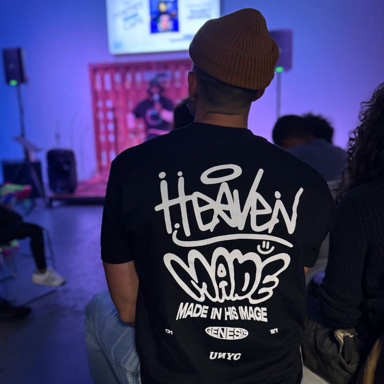 Person wearing a black t-shirt with white graffiti-style text, a beanie, in a dimly lit room with a stage and screen in the background.