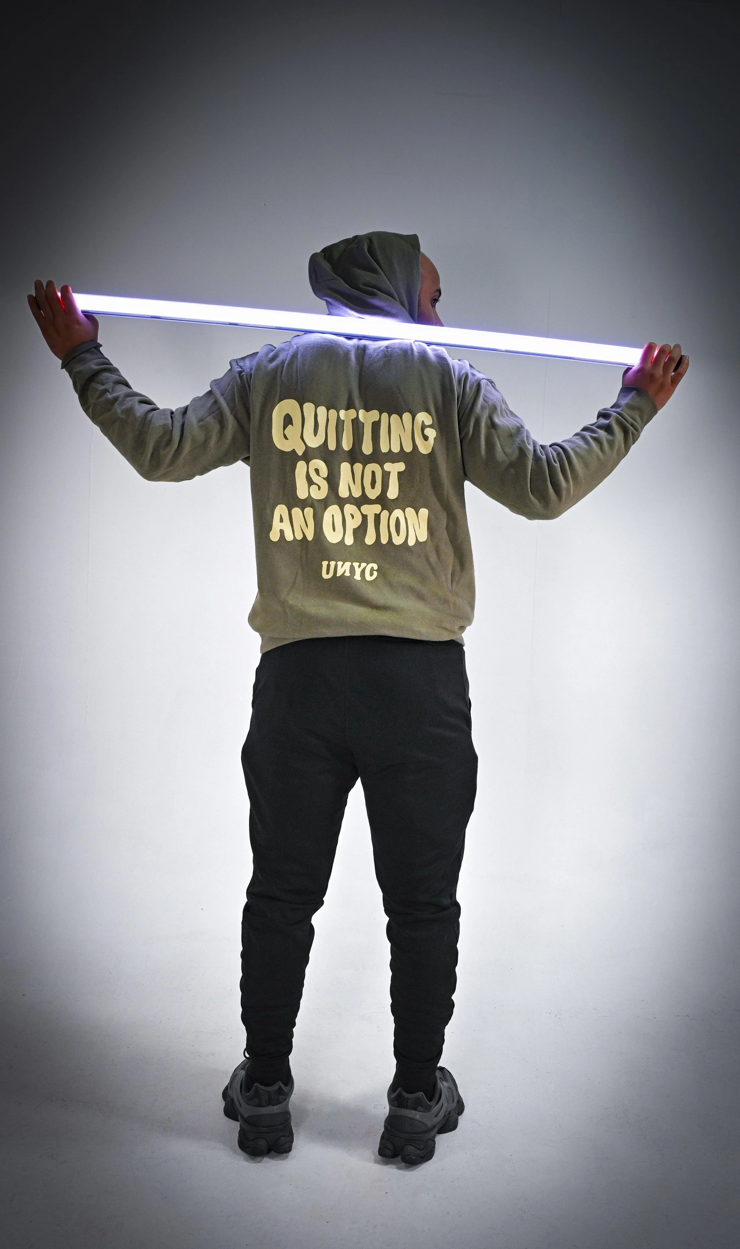 Person standing with back facing the camera, holding a horizontal neon light, wearing a hoodie with the text 'Quitting is not an option' on the back.