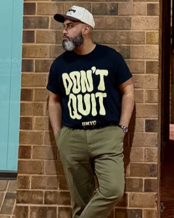Man with gray beard wearing a white cap, black t-shirt with 'DON'T QUIT' slogan, and khaki pants, standing against a brick wall