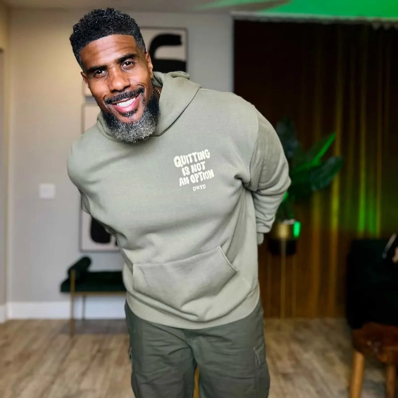 A man with dark hair and a beard smiling, wearing an olive green hoodie that says "Quitting is not an option" in white letters, and standing in a room with wooden floors and modern decor.