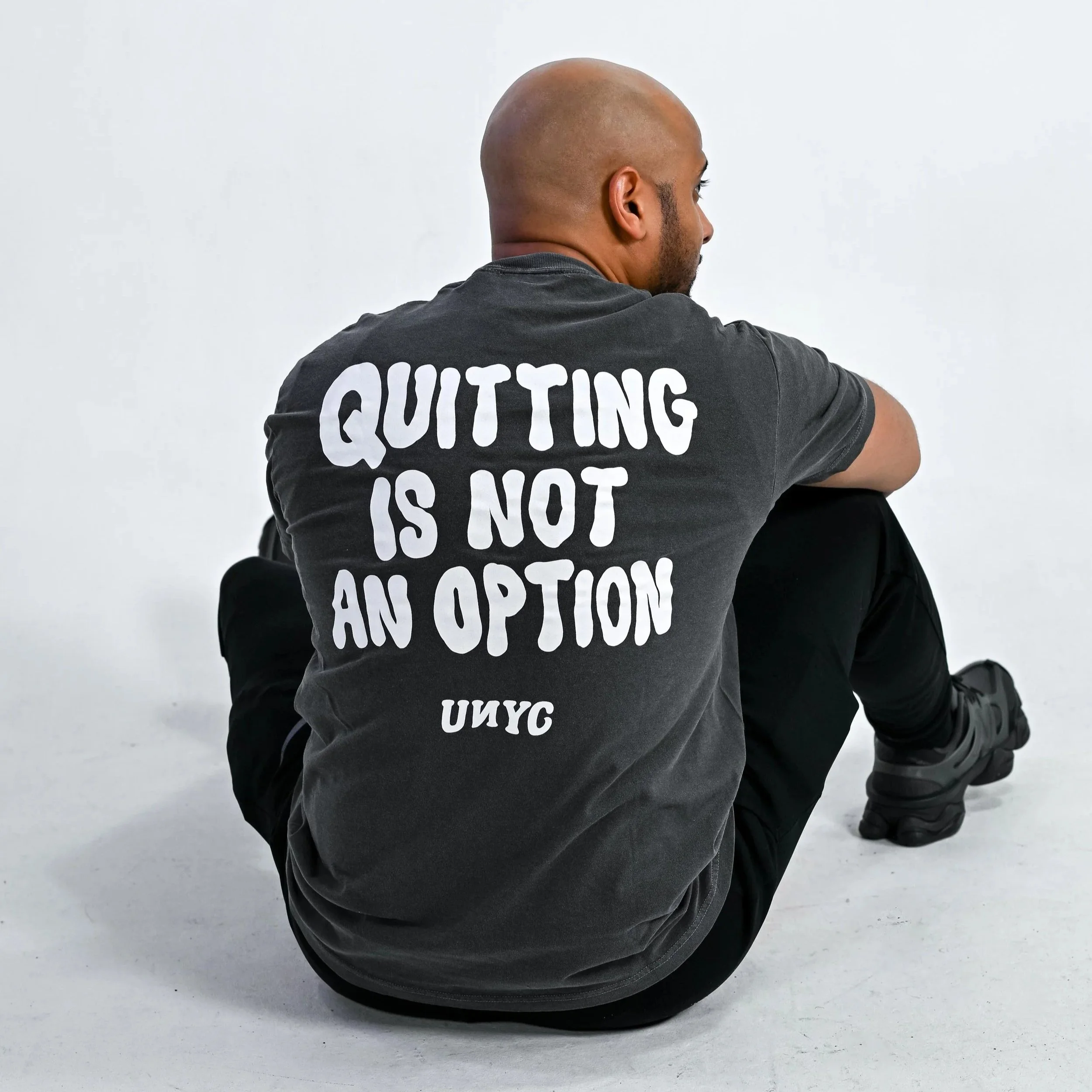 Person sitting with back facing camera, wearing a dark t-shirt with white text that says 'QUITTING IS NOT AN OPTION', in front of a white background.