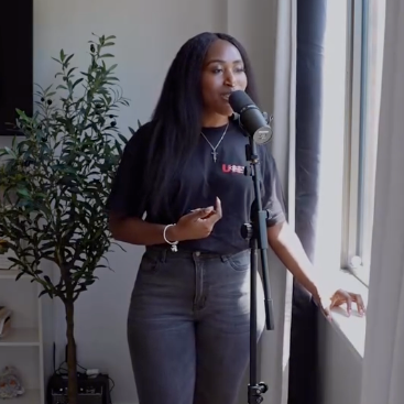 Woman speaking into a microphone during a recording session, standing near a window with sunlight, in a room decorated with a potted plant.