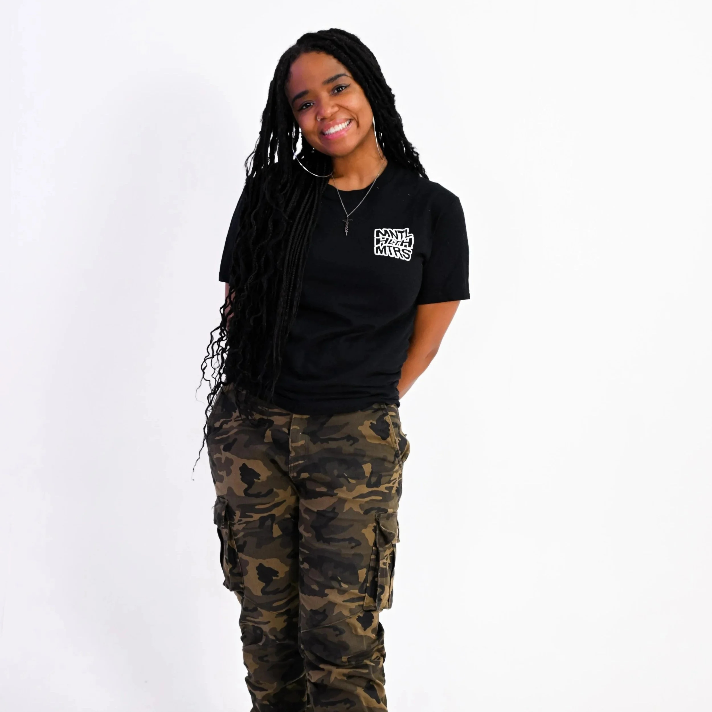 A young woman with long, braided black hair smiling slightly, wearing a black T-shirt with white text, hoop earrings, a necklace, and camouflage pants, standing against a plain white background.