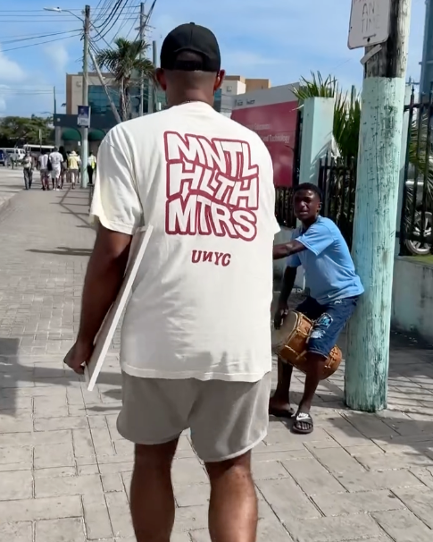 A man with a black cap and a cream T-shirt with maroon letters, walking on the sidewalk, with a man sitting on a curb holding a drum.