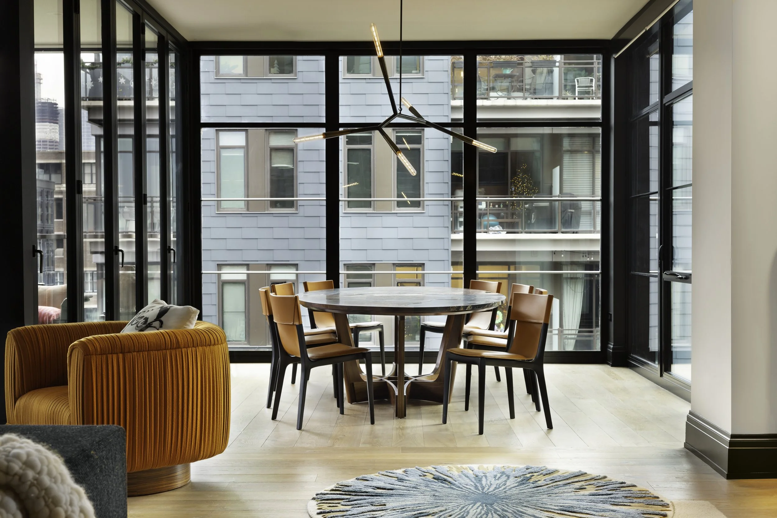 Modern dining room with a custom round walnut dining table and leather dining chairs, illuminated by a contemporary sculptural chandelier, set within a luxury urban loft featuring floor-to-ceiling black-framed windows and warm wood flooring.