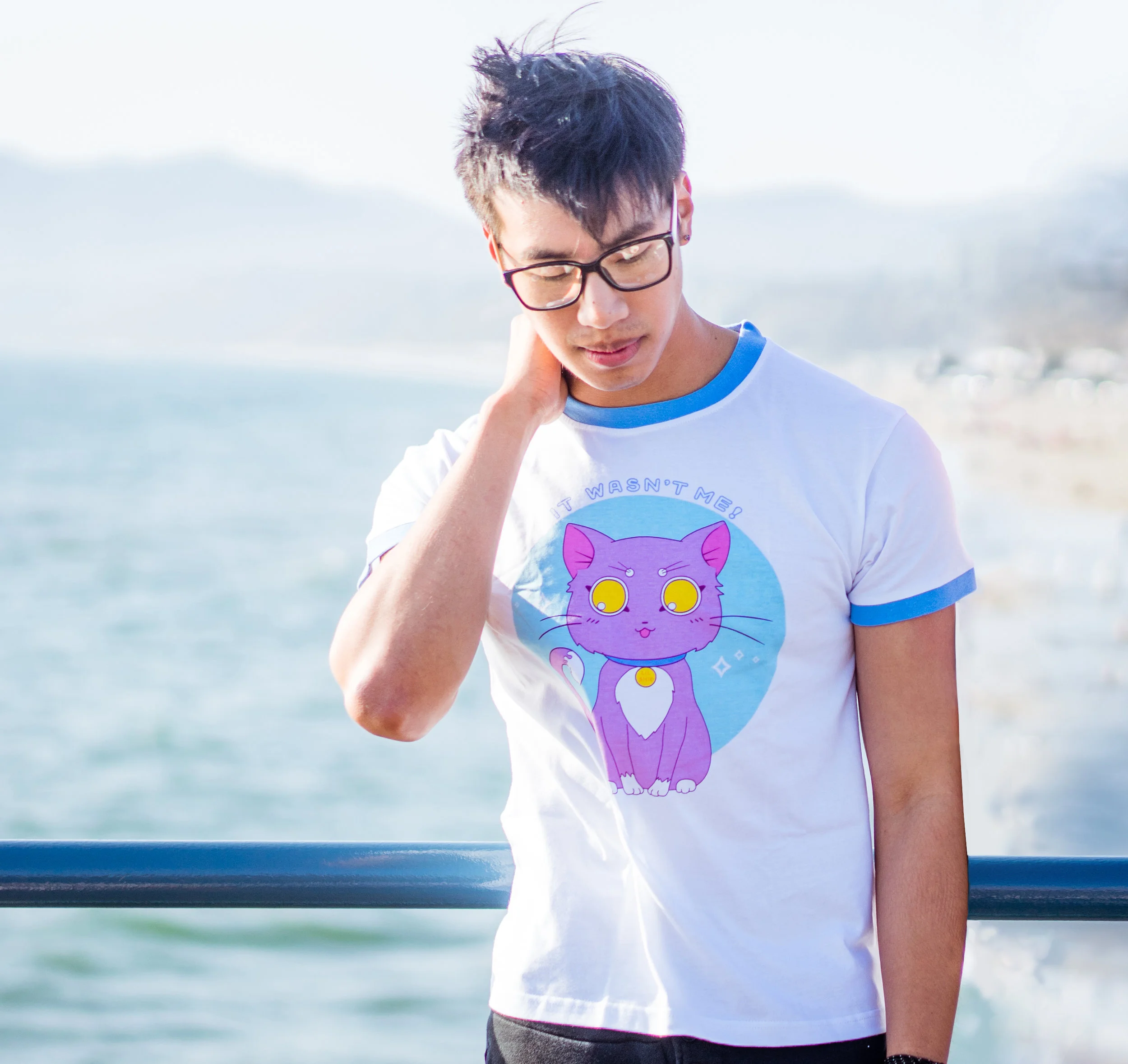 Young man with glasses and dark hair outdoors by the water, looking down, wearing a white t-shirt with a purple cartoon cat and the text 'It Wasn't Me'.