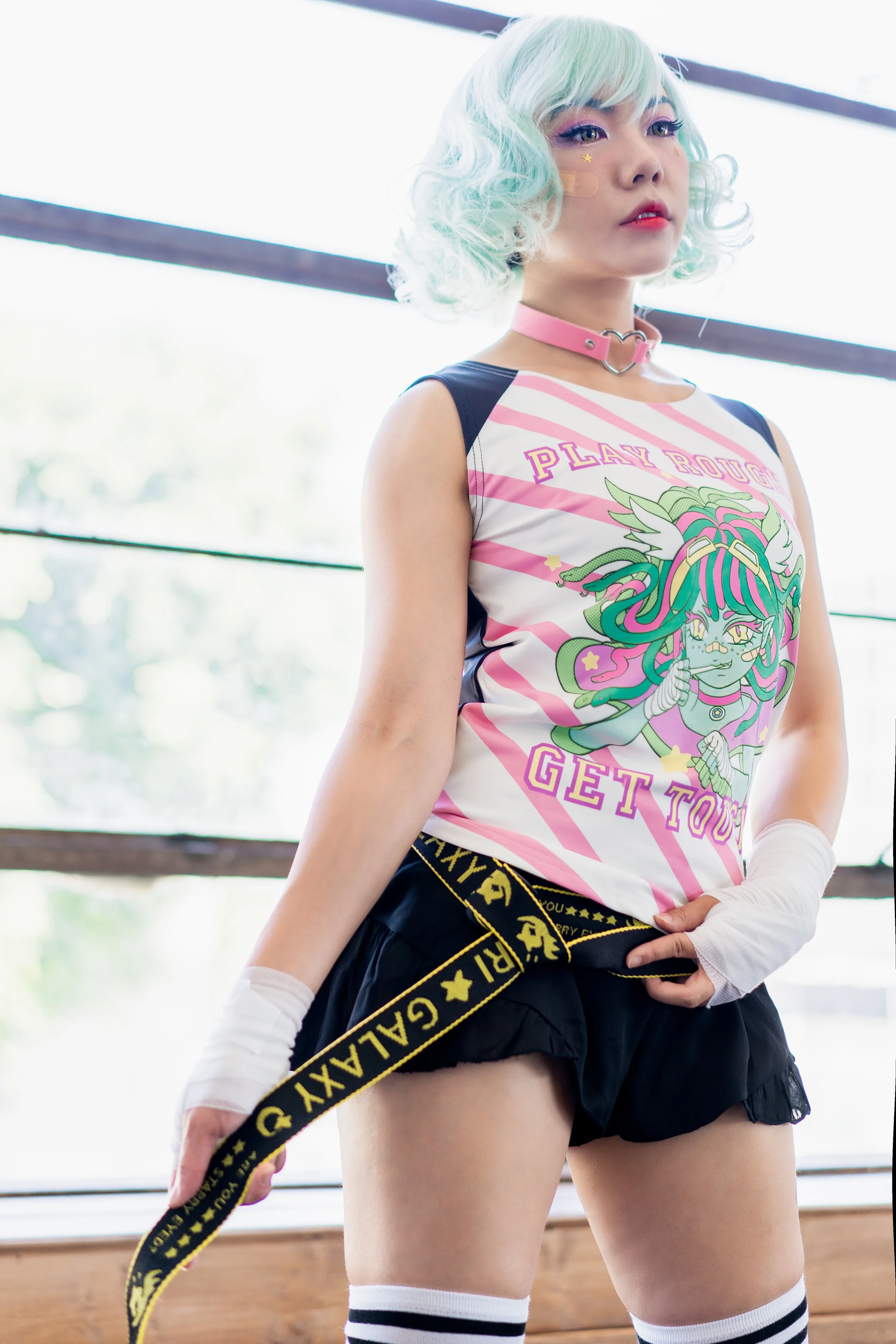 A woman with pastel mint green, curly hair and colorful makeup, wearing a graphic tank top, black shorts, striped knee-high socks, and a black and yellow belt, posing indoors near a large window.