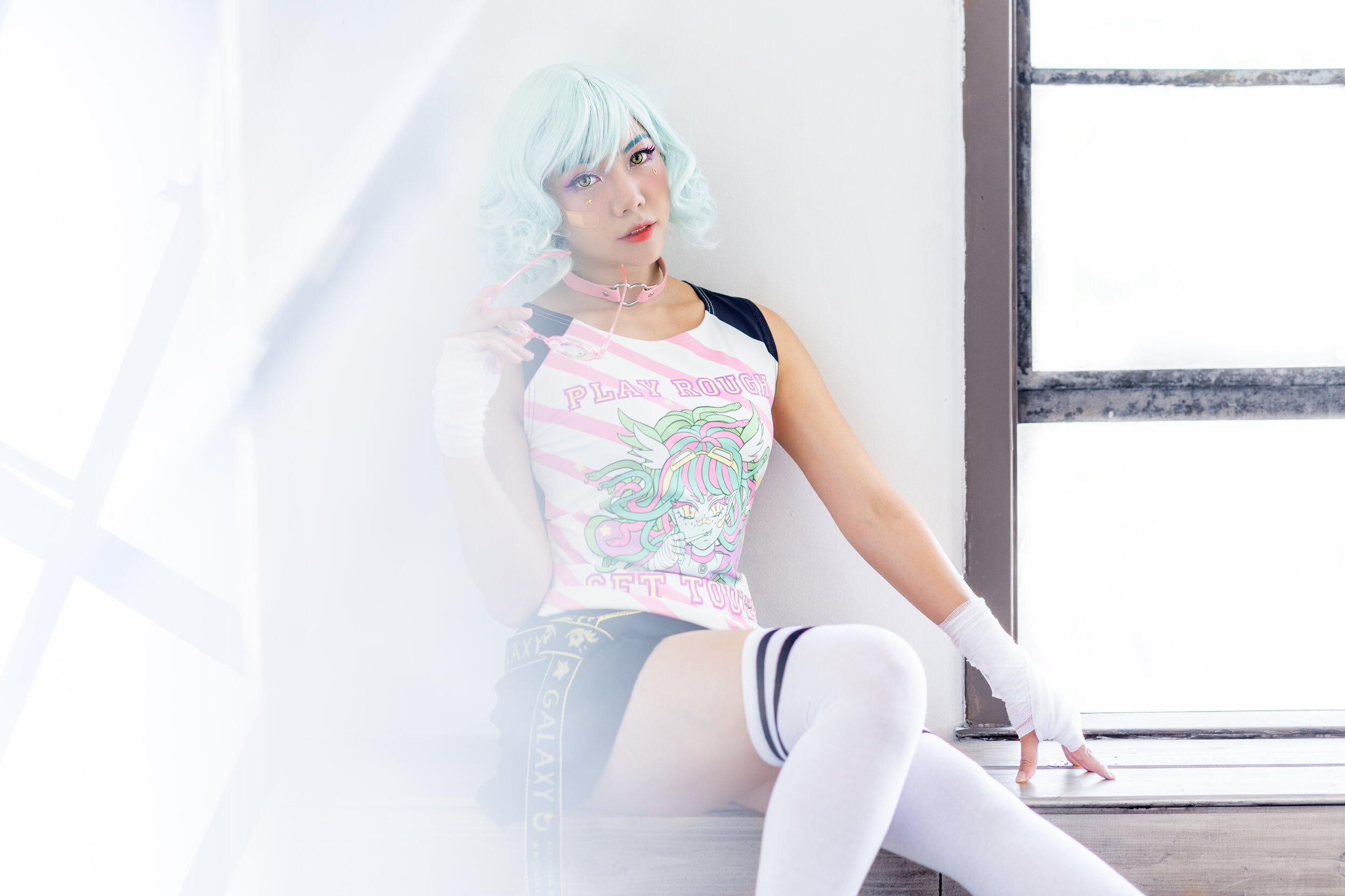 A young woman with pastel blue hair, wearing colorful anime-themed clothing, sitting on a windowsill with a bright, industrial-style window behind her.
