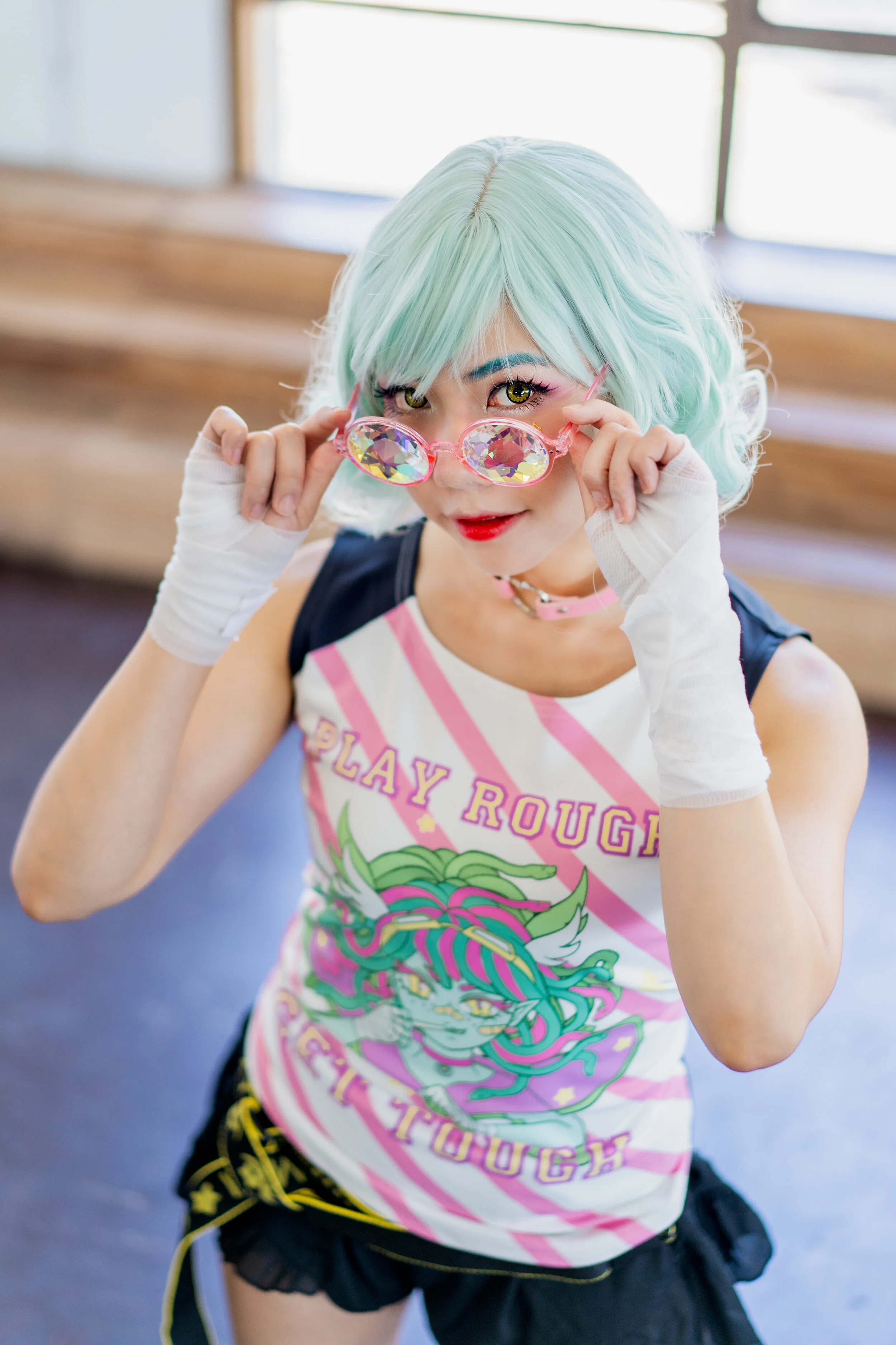 Person with light mint green hair, wearing a pink choker, colorful graphic sleeveless shirt, and black shorts, holding pink, jewel-encrusted glasses, in a room with wooden bleachers and large windows.