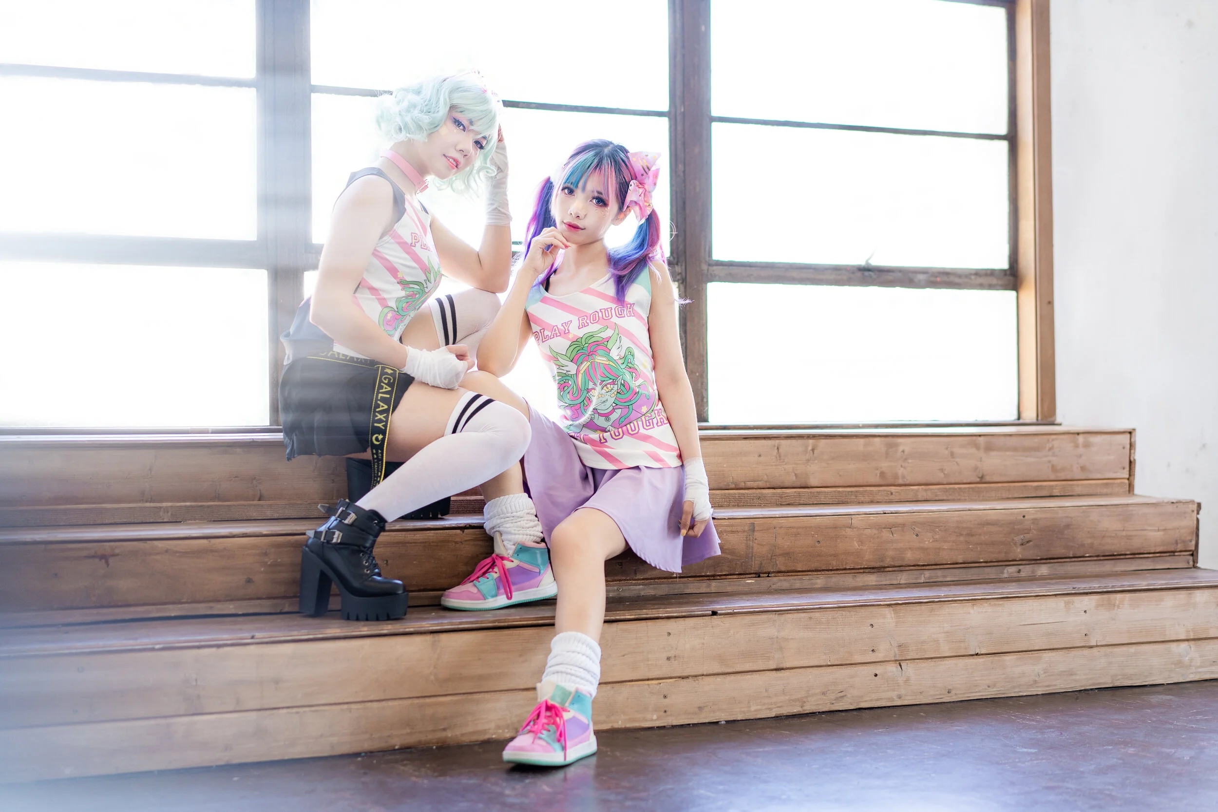 Two young women in colorful casual fashion sit on wooden steps in front of a large window with bright light, one wearing a sleeveless top, skirt, and platform boots, the other in a tank top, skirt, and sneakers, both with vibrant hairstyles and acces