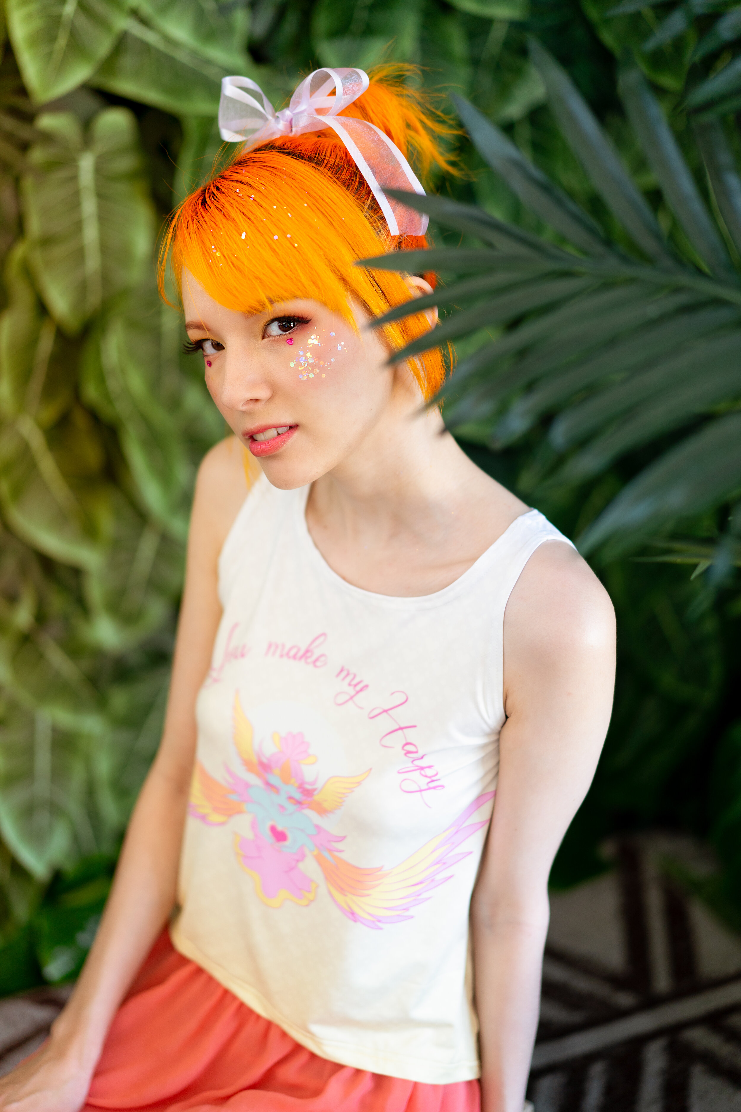 A young woman with bright orange hair styled with a bow and glitter, wearing a sleeveless off-white top with a flying pink and yellow unicorn design, and a coral skirt, standing among green tropical plants.