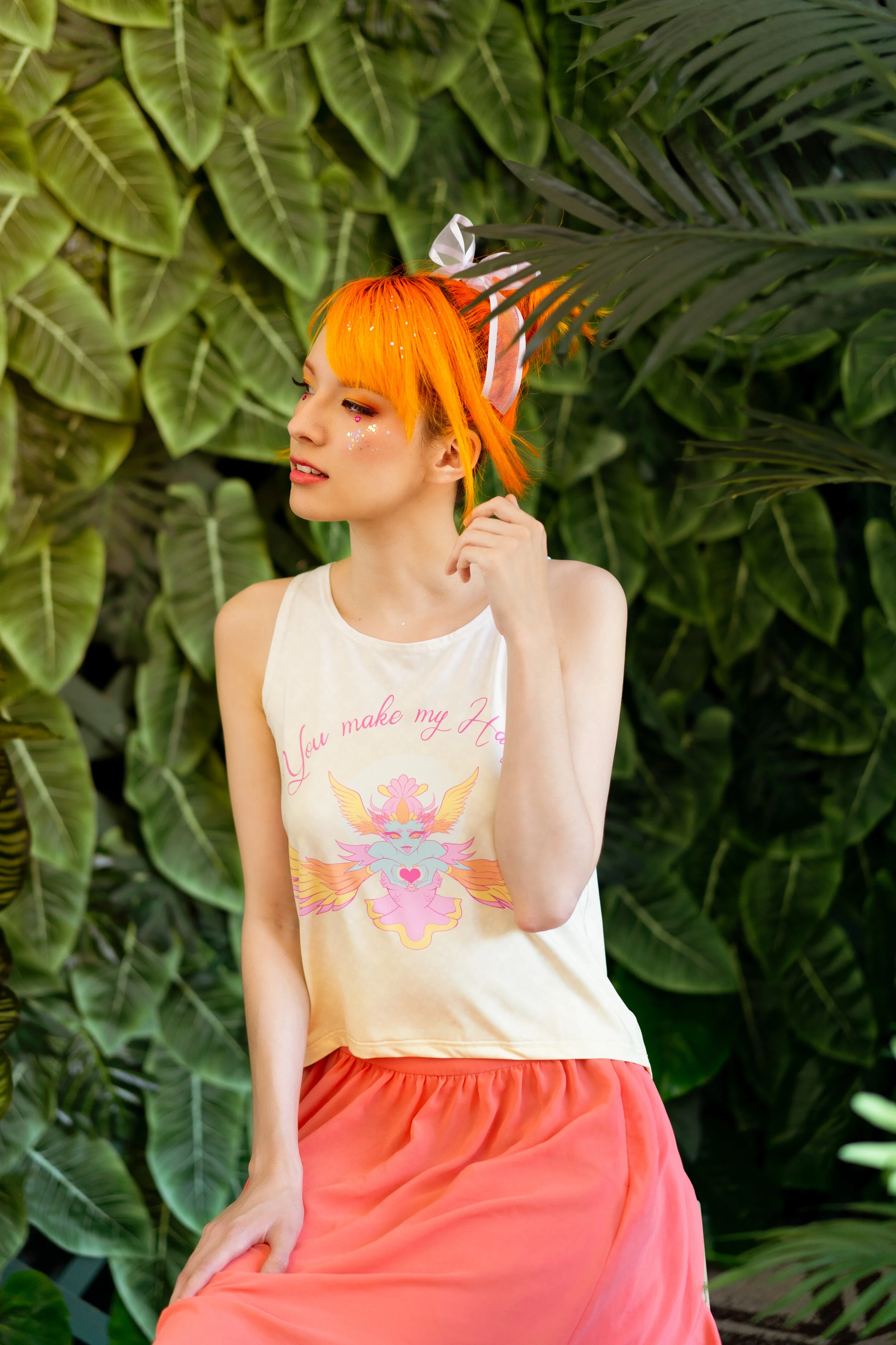 A woman with bright orange hair and glitter on her face, wearing a white tank top with a pink unicorn graphic and the text "You make my Heart"; she is sitting amidst a lush background of large green tropical leaves.