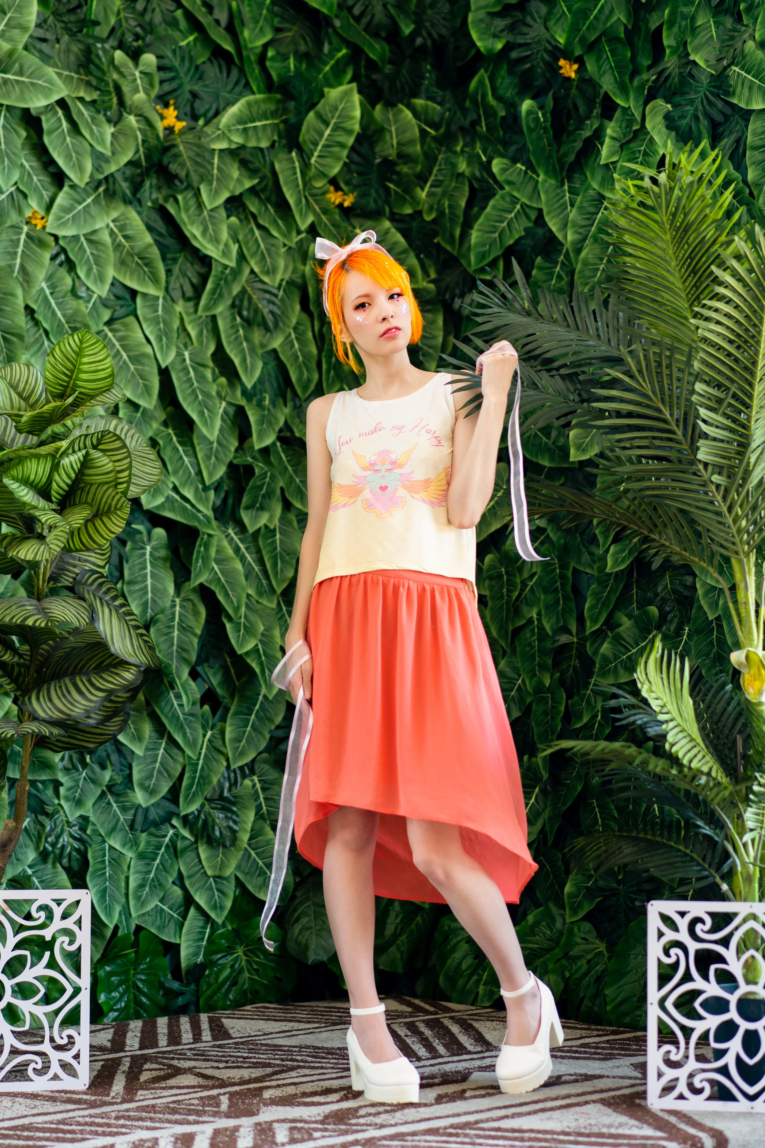 Young woman with orange hair and white bow, wearing a sleeveless graphic top and a coral skirt, standing in front of a lush green leafy background, holding a ribbon, wearing white platform heels.
