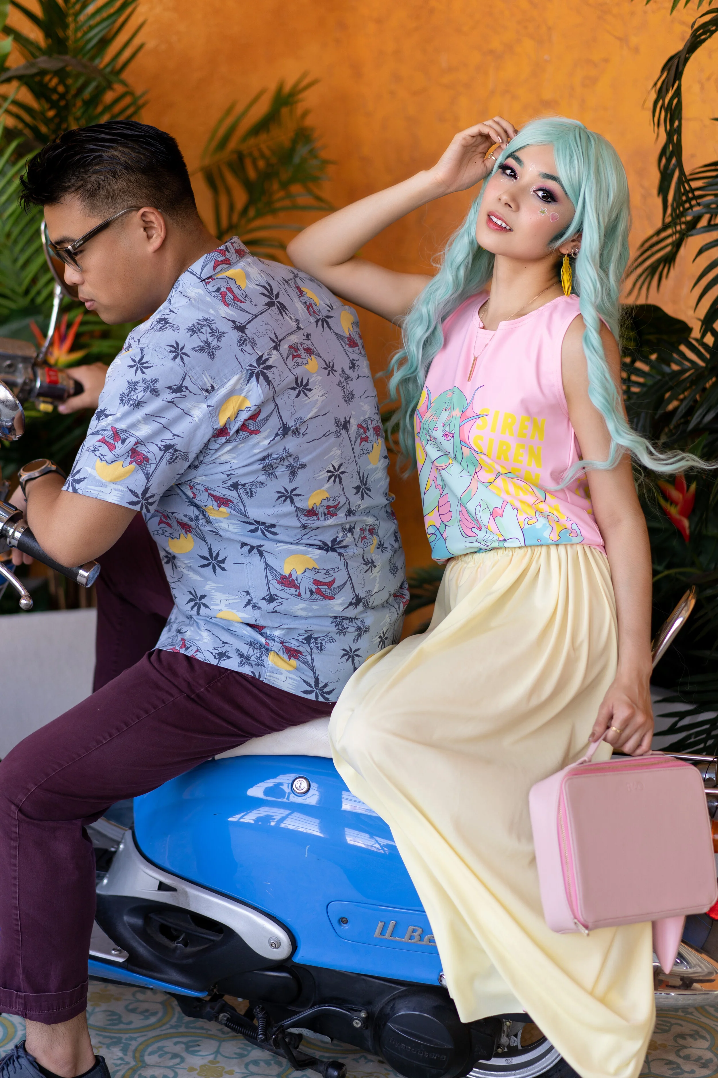 A woman with long teal hair, wearing a pink tank top with a mermaid design and yellow skirt, sitting on a blue scooter, with a pink purse hanging from her hand. A man with black hair, glasses, and a Hawaiian shirt, is sitting on the scooter with his 