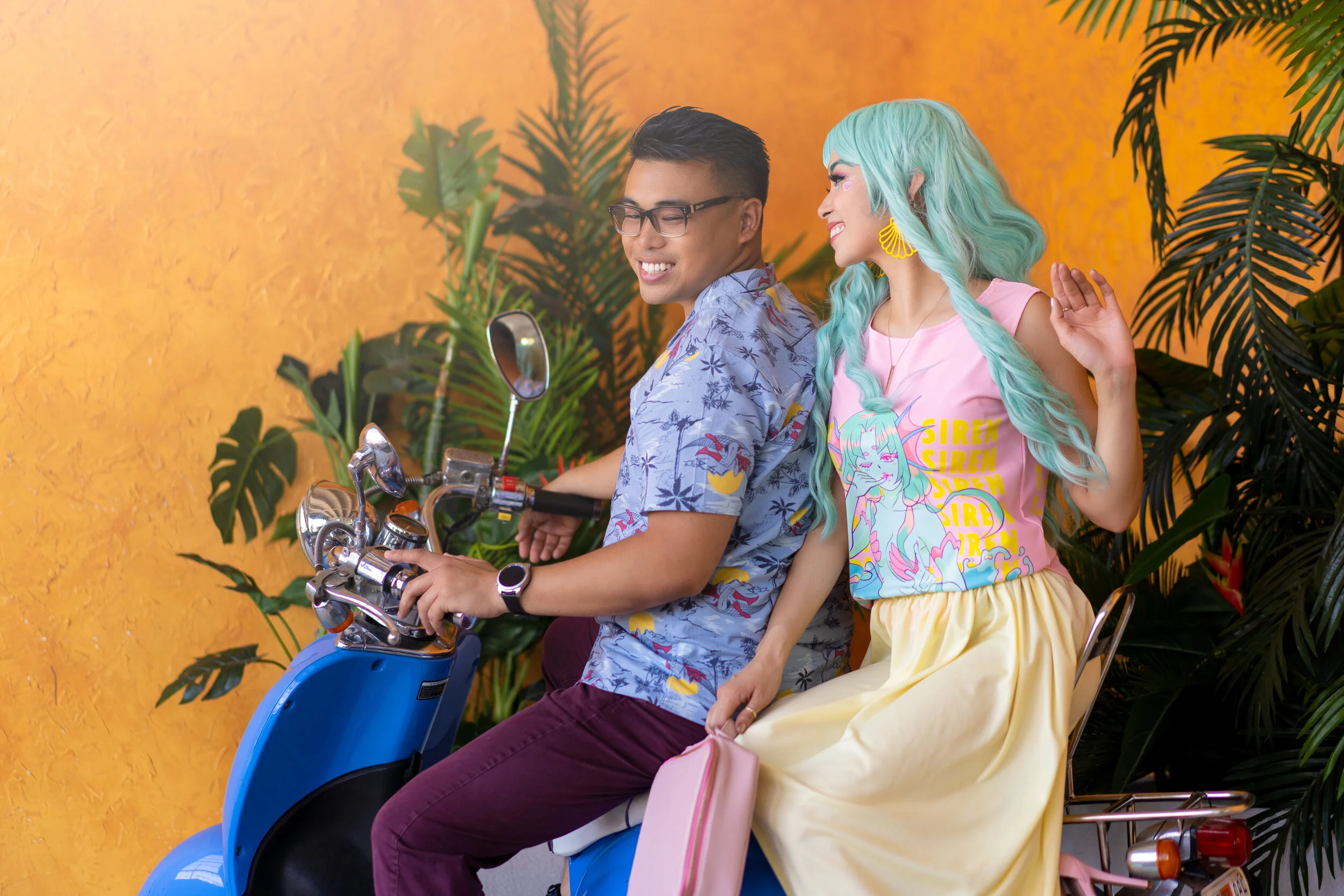 A young man and woman riding a blue scooter, with the woman sitting behind and holding onto the man. They are smiling and have colorful, tropical-themed clothes. The background features orange wall and lush green plants.