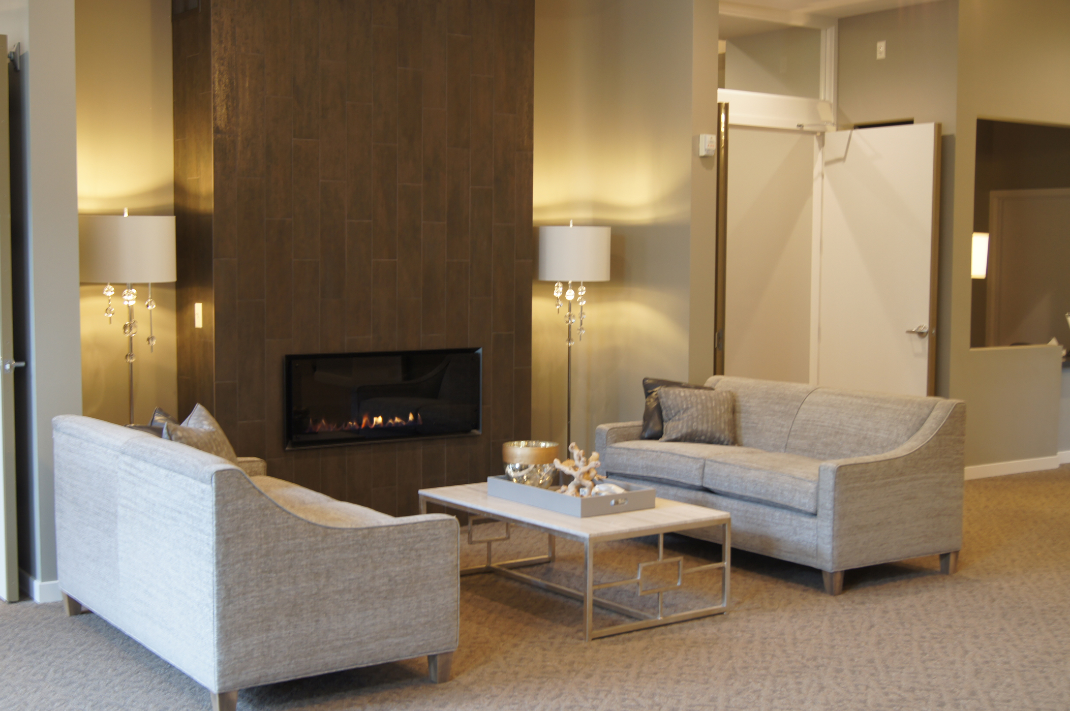AFTER: MAIN ENTRANCE LEFT SIDE ALL THE DARK OAK PANELING WAS REMOVED. A TWO-SIDED FIREPLACE WAS ADDED NOT ONLY AS A FOCAL POINT OFF THE MAIN ENTRANCE, BUT FOR COMFORT AND WARMTH ALSO ALLOWING AN AMAZING TILE TO BE SHOWCASED. NEW FURNITURE, LIGHTING AND ACCESSORIES ALL COORDINATE WITH THE GREY COLOR PALETTE ACCENTED WITH METALLIC + NATURAL FINISHES