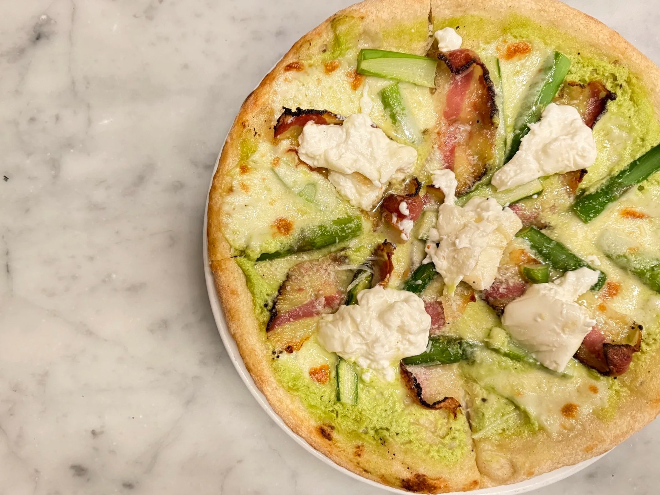 Pizza Special - Thin and round-shaped pizza, a green sauce as a base, based on asparagus, topped with mozzarella burrata, crispy guanciale, and tips of asparagus.
