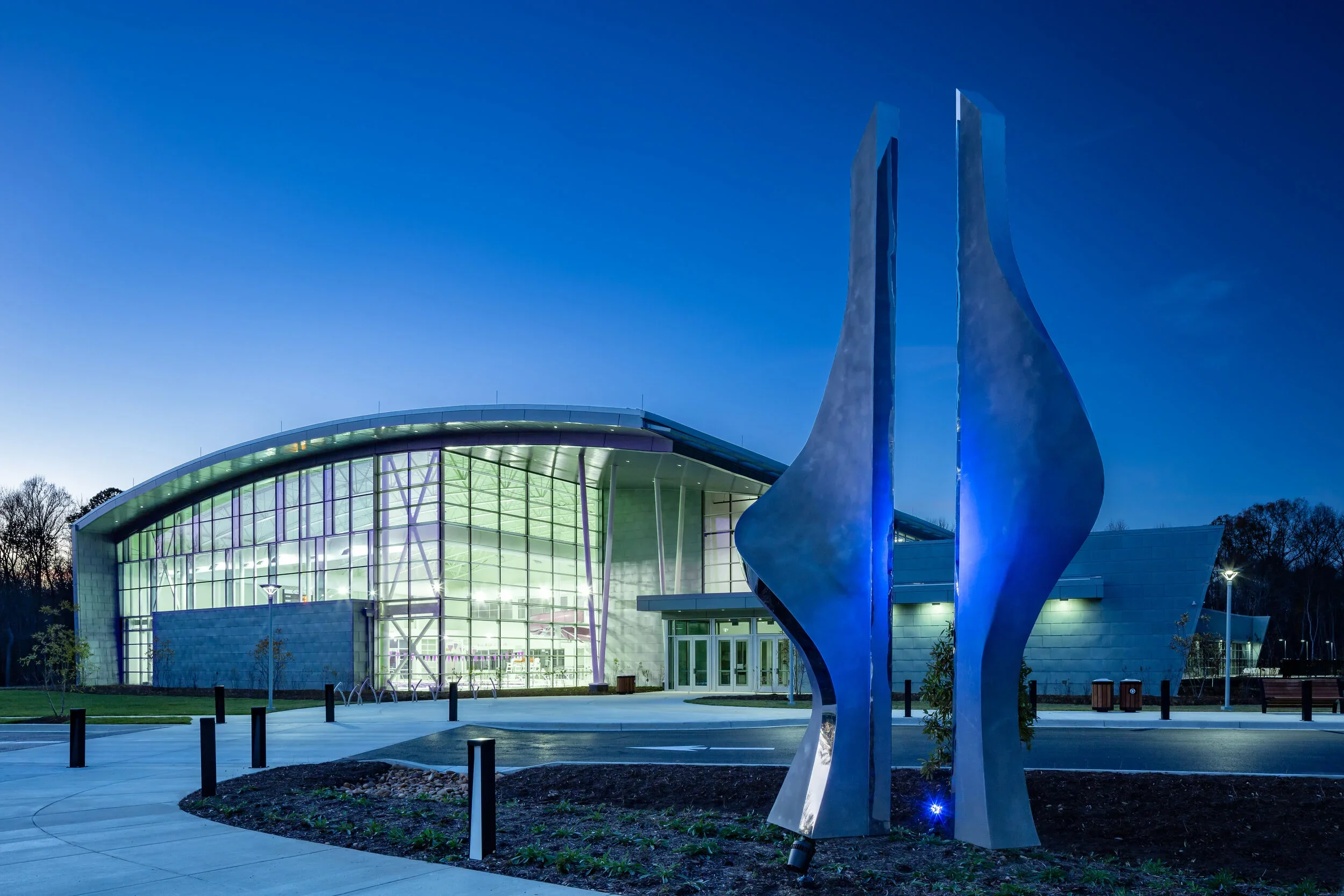 SAARC | Nighttime view of a modern building with glass front, illuminated inside, and artistic metal sculptures in front, surrounded by trees and a paved walkway.