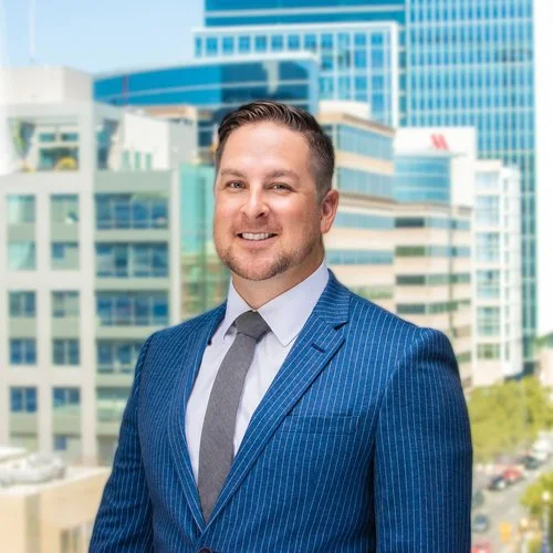A smiling man in a blue pinstripe suit with a gray tie standing outdoors in front of modern office buildings.
