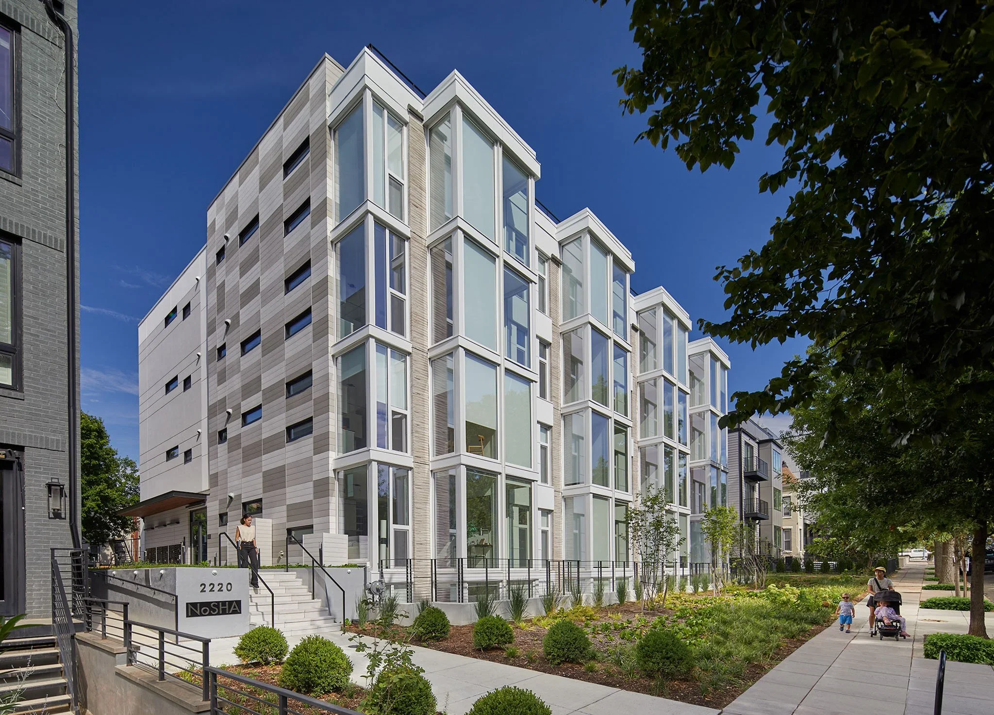 Modern multi-story residential building with glass balconies, surrounded by trees and a landscaped yard, with people walking on the sidewalk.