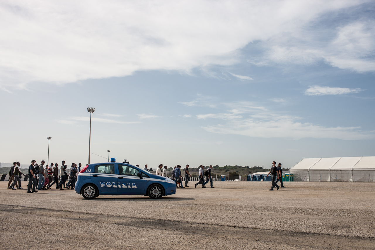Italian Police escort newly arriving migrants from the port where they docked to a nearby holding area. While most know the majority of at least the Syrian, Palestinian, and Eritrean refugees will escape within the following 48 hours, authorities still carry along with a certain song and dance.