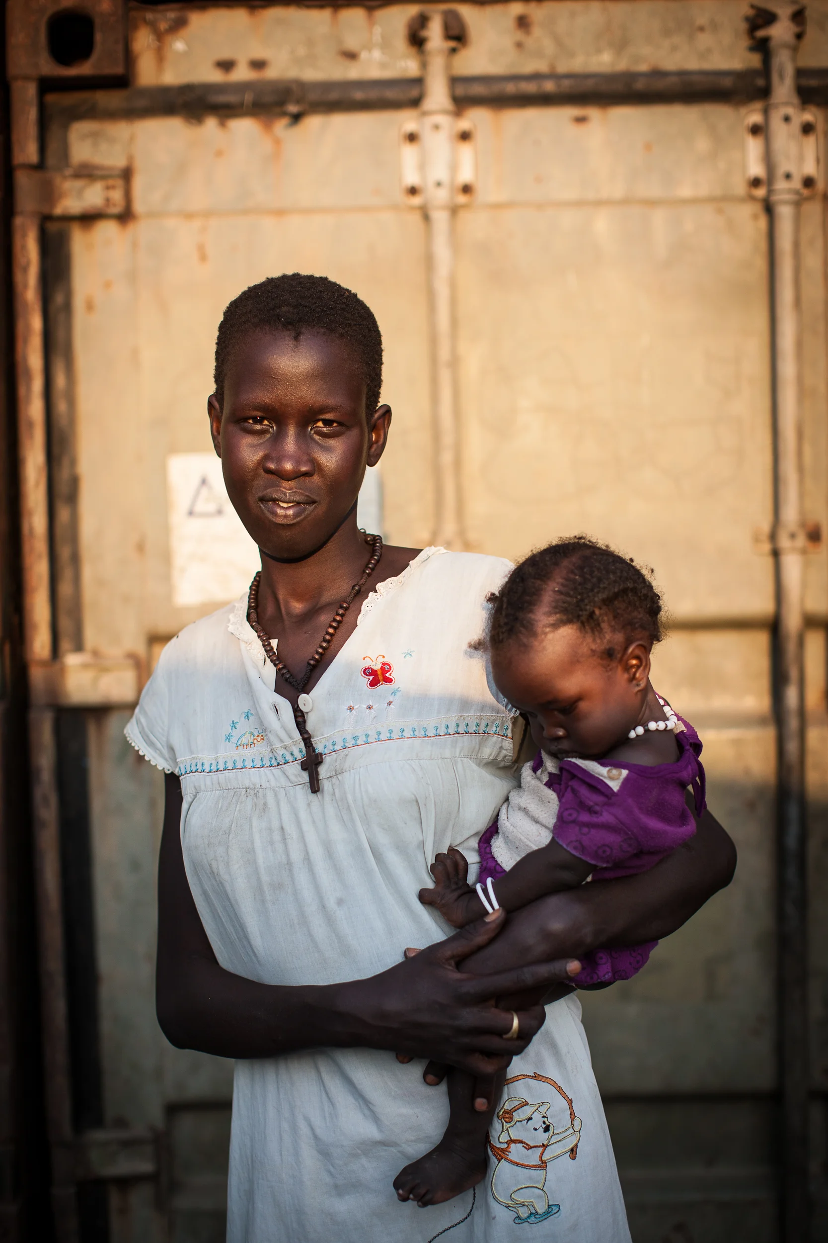 Rebecca, 18 stands with her 6-month old daughter, Gisma outside the shipping container where they live in the Malakal PoC.