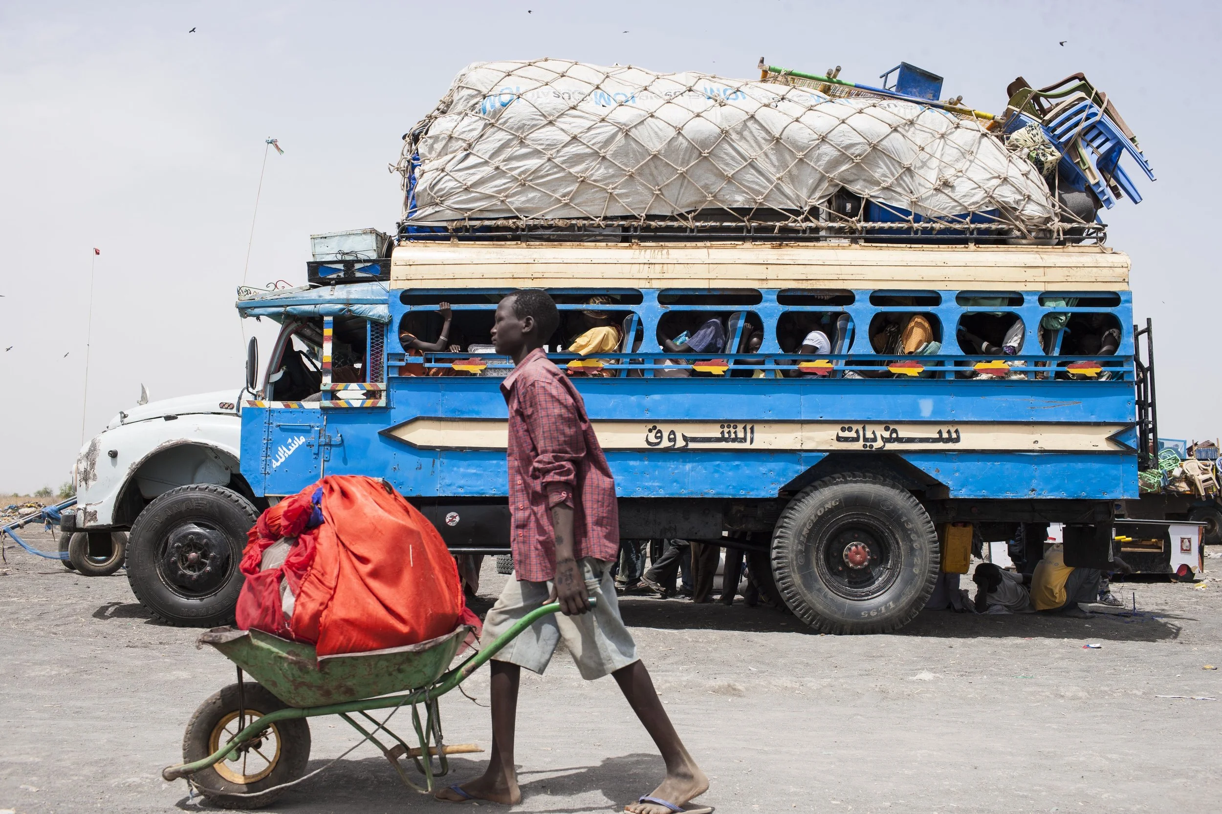 A boy pushes a wheelbarrow past the fully-loaded bus.