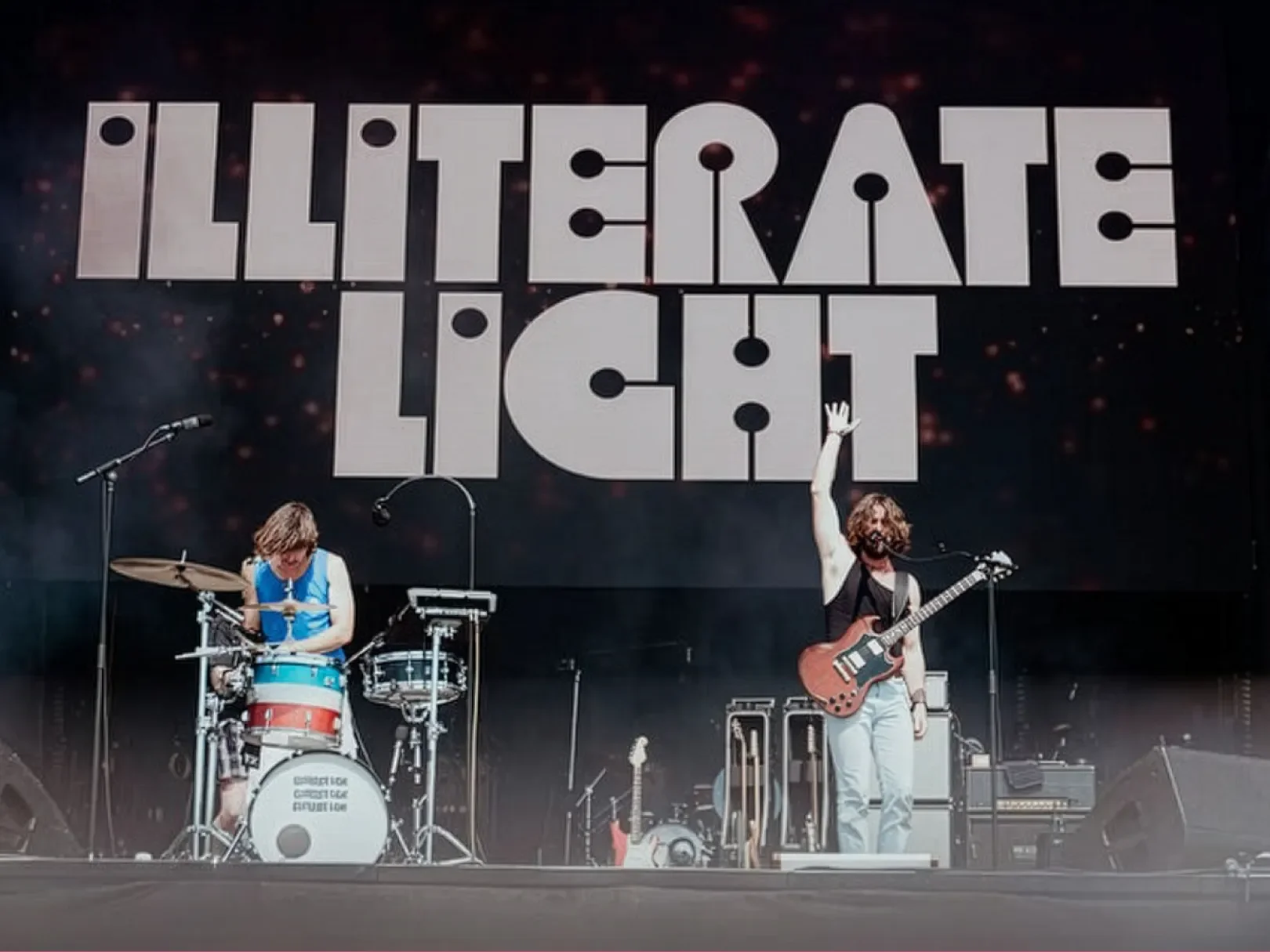 A band performing on stage with a large sign above them reading 'ILLITERATE LIGHT'. The drummer and guitarist are playing, with the guitarist raising one hand.
