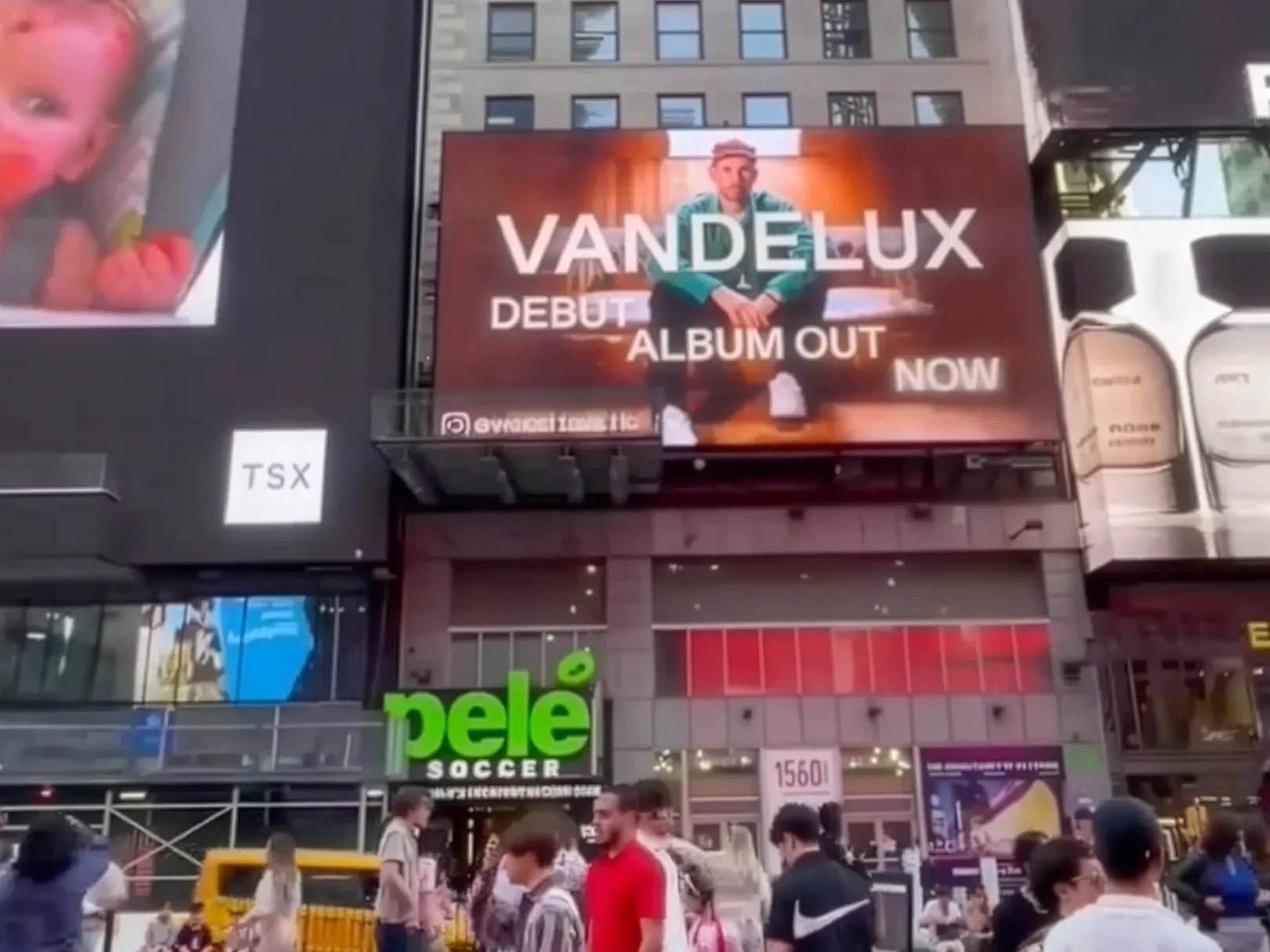 A busy city street with a large digital billboard advertising Vandelux's debut album out now, and multiple people walking on the sidewalk.