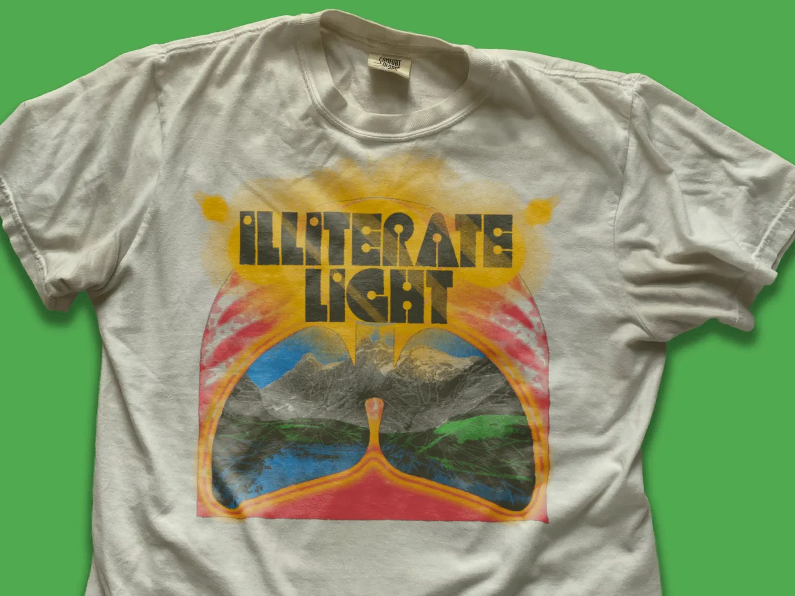 White t-shirt with a colorful graphic of mountains, lakes, and a sunrise, with the words 'ILLITERATE LIGHT' printed on top.