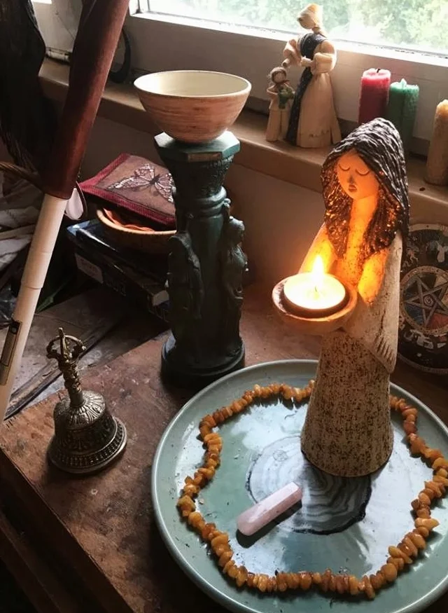Image by Arie Farnam - An altar for the goddess Brigid with a stature holding a lit candle surrounded by a string of amber beads and a rose quartz crystal in the foreground. In the background there is a pedestal with goddess carvings and an offering bowl at the top. Candles and a corn-husk doll sit on a window sill. To the left a white cane with a brown wood handle leans against the altar over a brass bell. 
